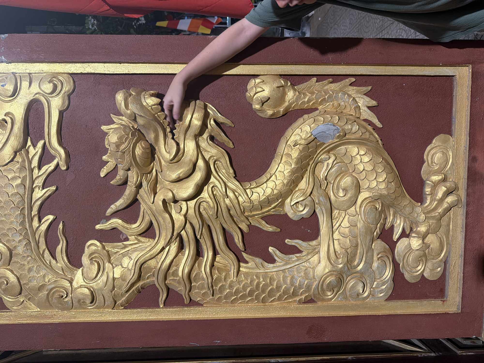 A close-up shot captures a detailed golden dragon relief on a maroon wall. A person's left arm and hand are visible, with fingers gently touching the dragon's snout, suggesting interaction with the artwork. The dragon is intricately carved with scales, flowing mane, and fierce facial features. The surrounding wall appears to be part of a building or structure, possibly a temple or monument. In the background, hints of a red flag and another flag with horizontal stripes of orange, yellow, and white can be seen, suggesting a cultural or religious setting. The lighting indicates it might be daytime, though shadows suggest an overcast condition or late afternoon. The image is taken in Đà Nẵng, Vietnam, a city known for its cultural heritage and architectural landmarks.