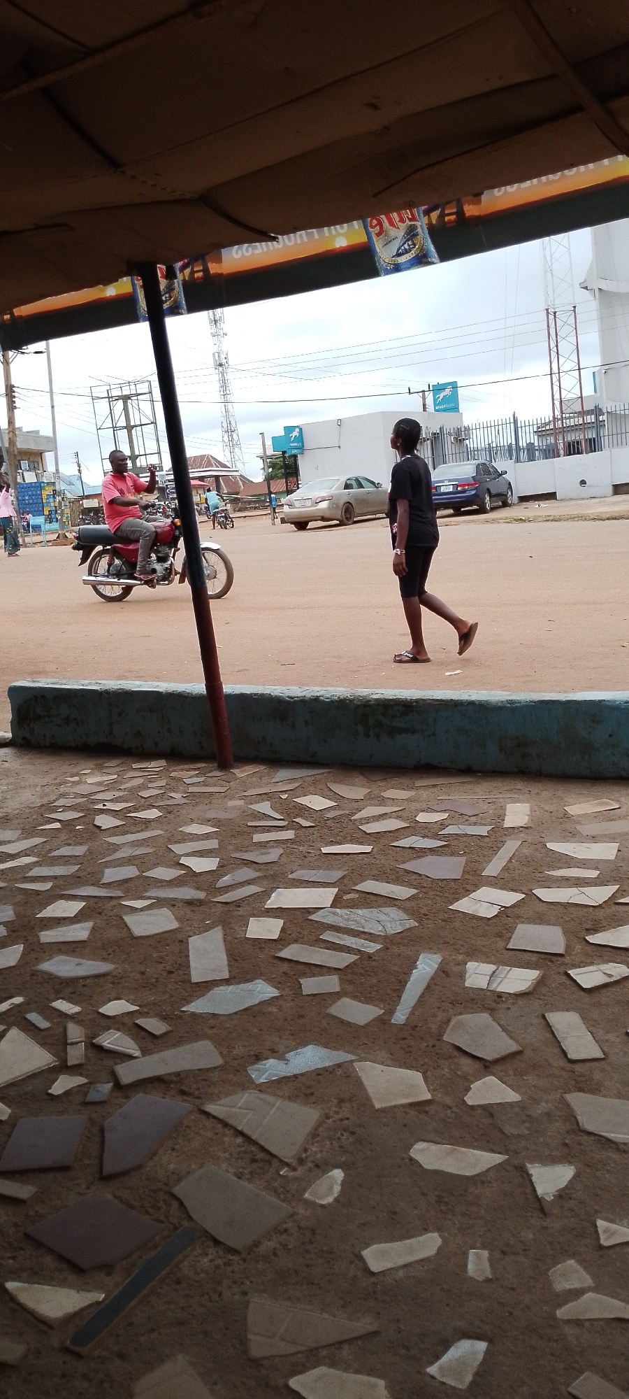 The image depicts a street scene in Gboko, Nigeria, under an overcast sky. In the foreground, a mosaic-tiled ground is visible, with broken pieces of tiles embedded in a darker material. A turquoise-colored concrete barrier runs horizontally across the middle of the frame, with a red cylindrical pole rising from it on the left.
Beyond the barrier, a dirt road is seen with vehicles and people. On the left, a man wearing a pink t-shirt and dark trousers rides a red motorcycle, holding up his left hand as if to wave or signal. To his right, a young woman with dark hair, wearing a black t-shirt and black shorts, walks away from the camera towards the right side of the image. She is wearing flip-flops.
In the background, several cars are present on the road, including a silver sedan and a dark blue sedan. A white building with a fence and some signage, possibly a bank, is visible behind the cars. A cellular tower stands tall in the distance. The general environment appears to be an urban or semi-urban area.