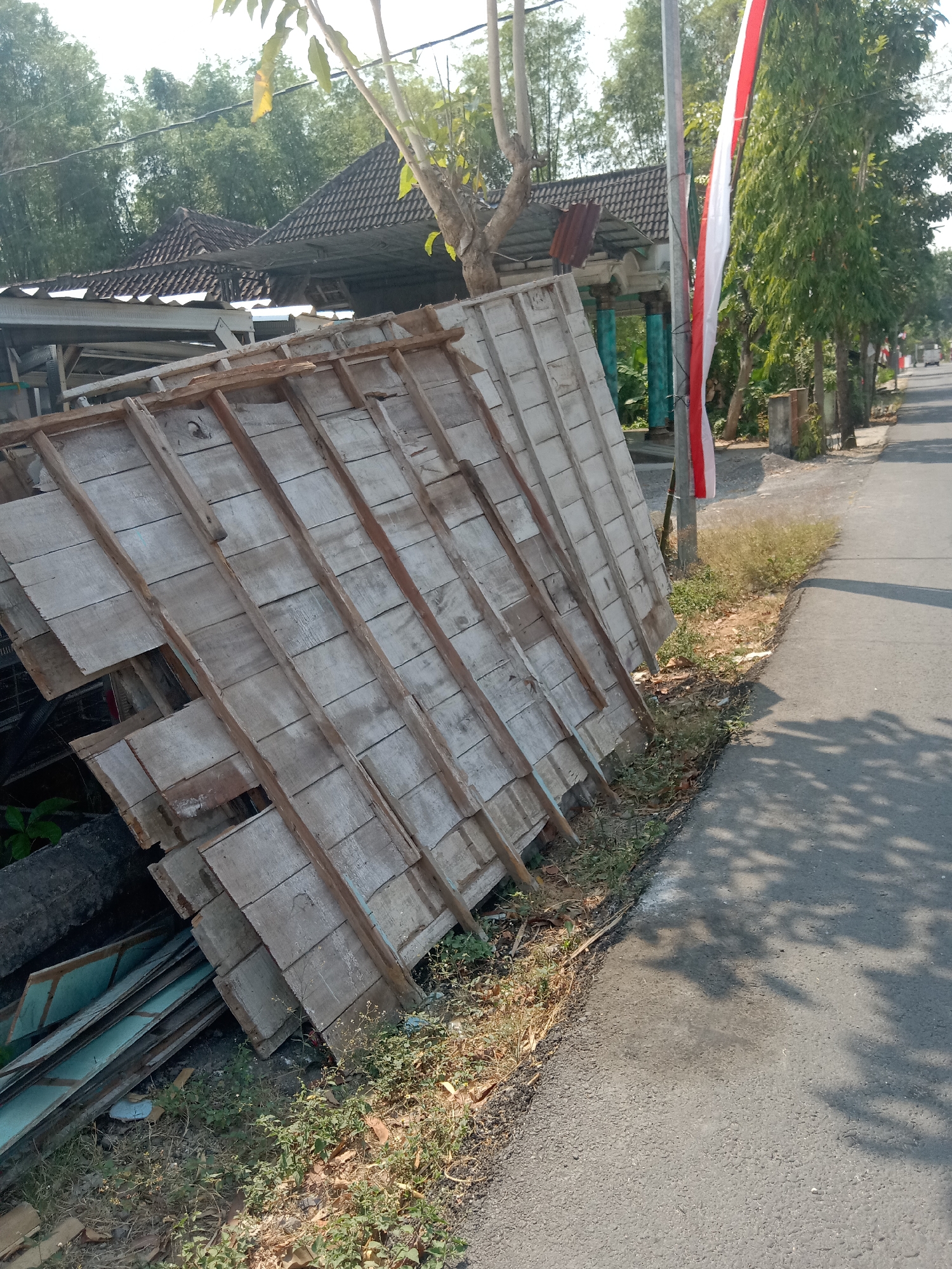 The image depicts an outdoor scene in Tegalkodo, Indonesia, on a sunny day. In the foreground, a pile of weathered wooden boards and planks is stacked diagonally against a pole. The wooden structure appears to be part of a dismantled construction or a collection of building materials. A red and white Indonesian flag is attached to the pole, fluttering gently. To the right of the wooden pile and pole is an asphalt road with shadows of trees cast across it. The road curves slightly to the right and extends into the distance, lined with trees on the right side. In the background, the roofs of several houses with tiled roofs are visible, surrounded by lush green trees. The overall environment suggests a residential or semi-rural area. There are no people visible in the image.