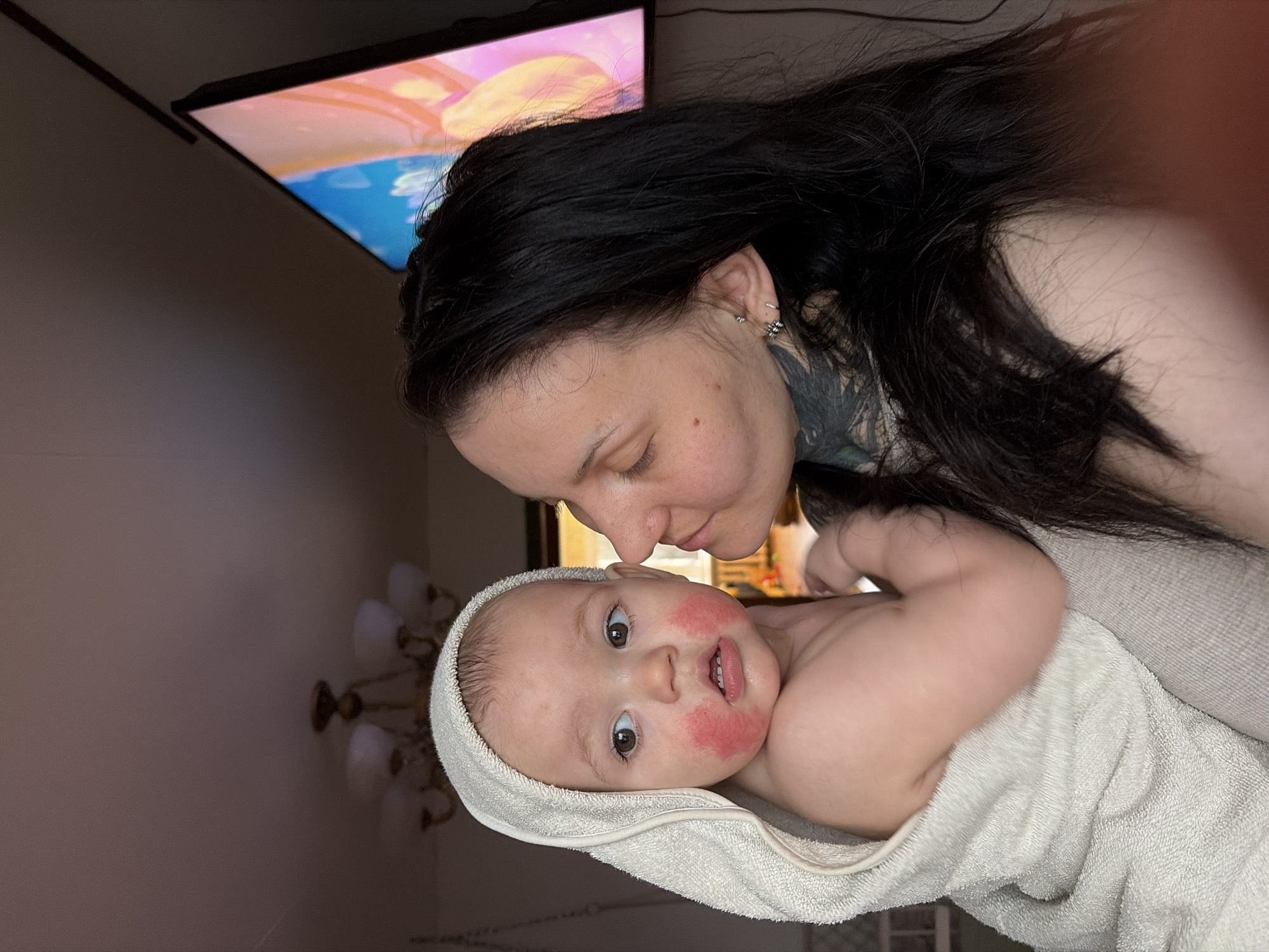 A woman with long, dark hair and a neck tattoo is close to a baby with redness on its cheeks. The baby is wrapped in a light-colored towel. They appear to be indoors, with a television and a chandelier visible in the background. The location is Chaikovsky, Russia.
