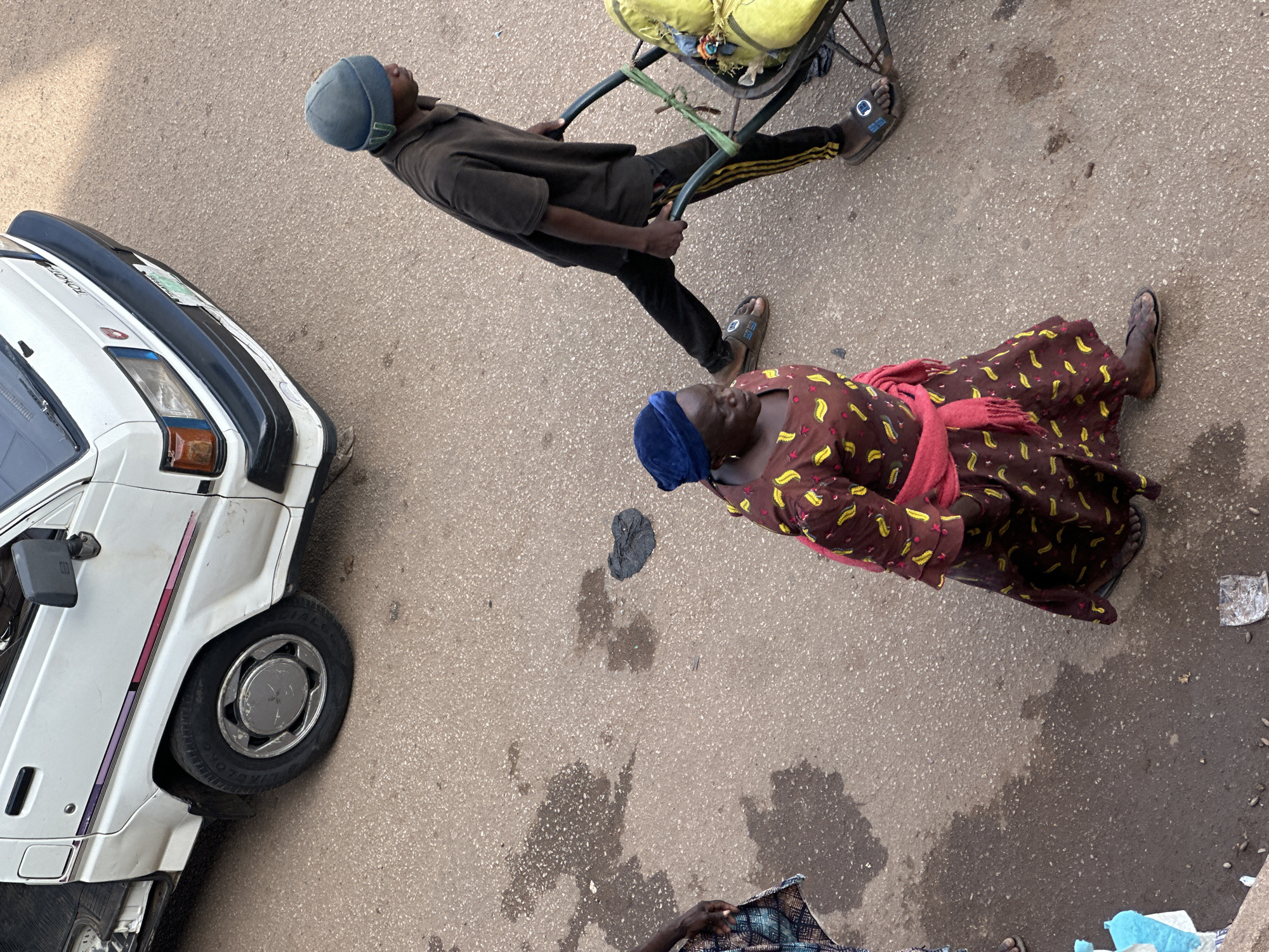 In Bukuru, Nigeria, an elevated, overhead shot captures a scene on a paved road with visible water stains and some debris. A white Toyota van, partially visible on the left, is parked on the road. In the foreground, a woman wearing a brown dress with yellow banana-like patterns and a blue headscarf is walking. She has a pink shawl draped around her shoulders. To her right, a young man wearing a dark t-shirt, dark pants with yellow stripes, and a blue beanie is pushing a wheelbarrow loaded with yellow sacks. His movement suggests he is walking away from the camera, following the woman. Another person, partially visible on the far left, is wearing a patterned skirt. The lighting indicates it is daytime, possibly late afternoon due to the shadows.