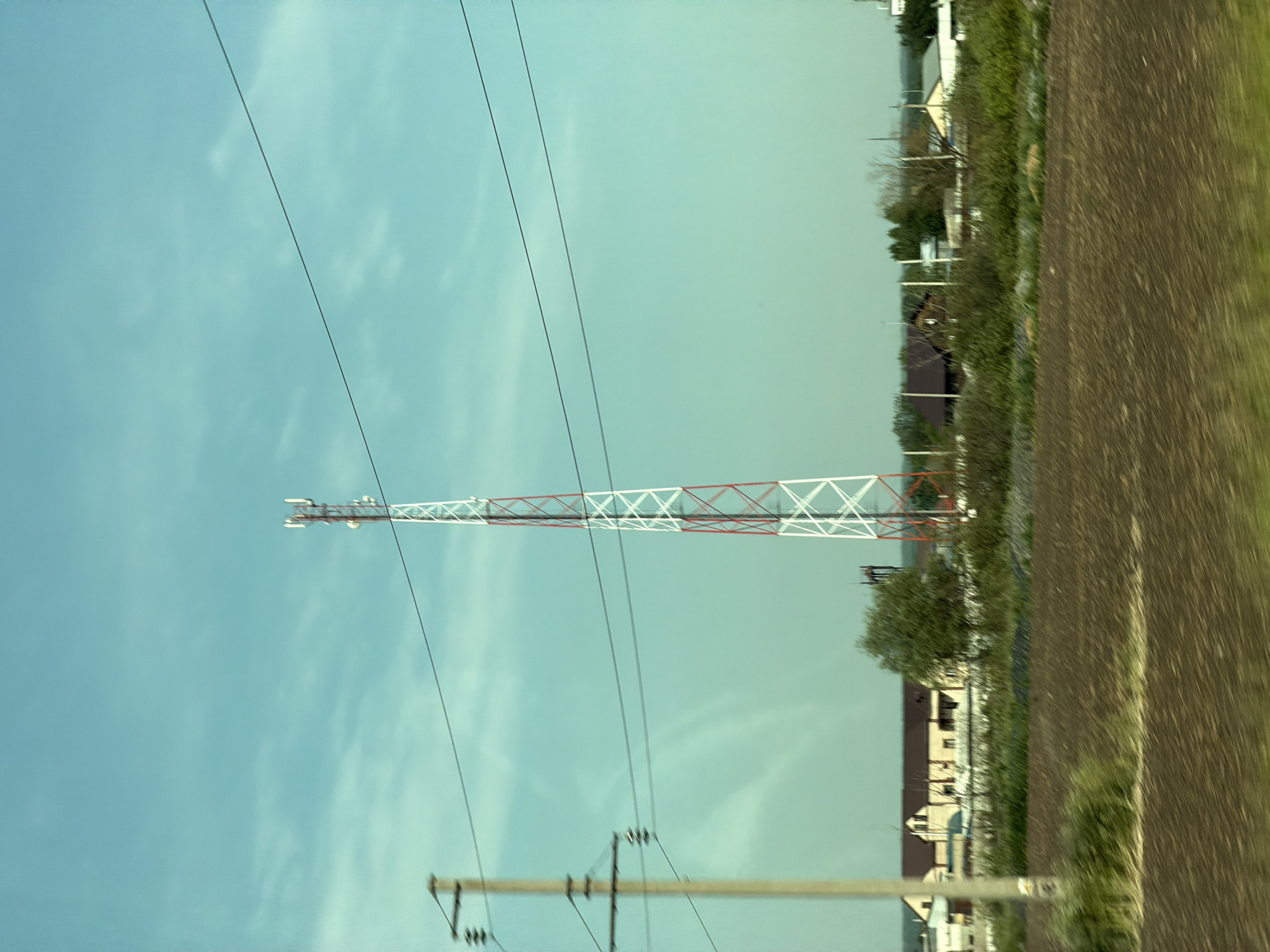 The image captures a cellular tower situated in the city of Evsino, Russia. The tower, painted white and red, is tall and slender, with multiple antennas at its apex. It stands against a backdrop of a clear, pale blue sky dotted with wisps of clouds. In the middle ground, several houses with dark roofs are visible, nestled amidst green vegetation and trees. A power line stretches across the frame, supported by a concrete pole on the left. The foreground consists of a ploughed field, its brown earth showing the marks of agricultural activity. The overall impression is of a rural or semi-rural setting on a bright, clear day.