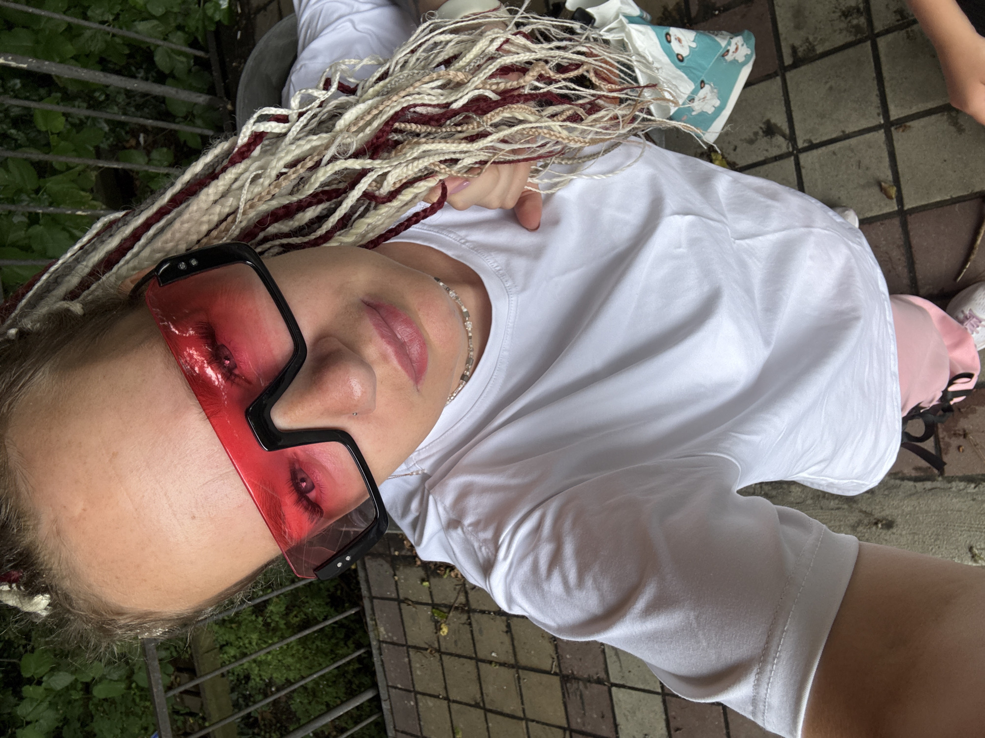 A young woman with blonde and burgundy dreadlocks, wearing large red-tinted sunglasses, a white t-shirt, and pink shorts, is taking a selfie outdoors. She is standing on a paved walkway made of square tiles, with a metal fence and green foliage visible in the background. The lighting suggests it is daytime.