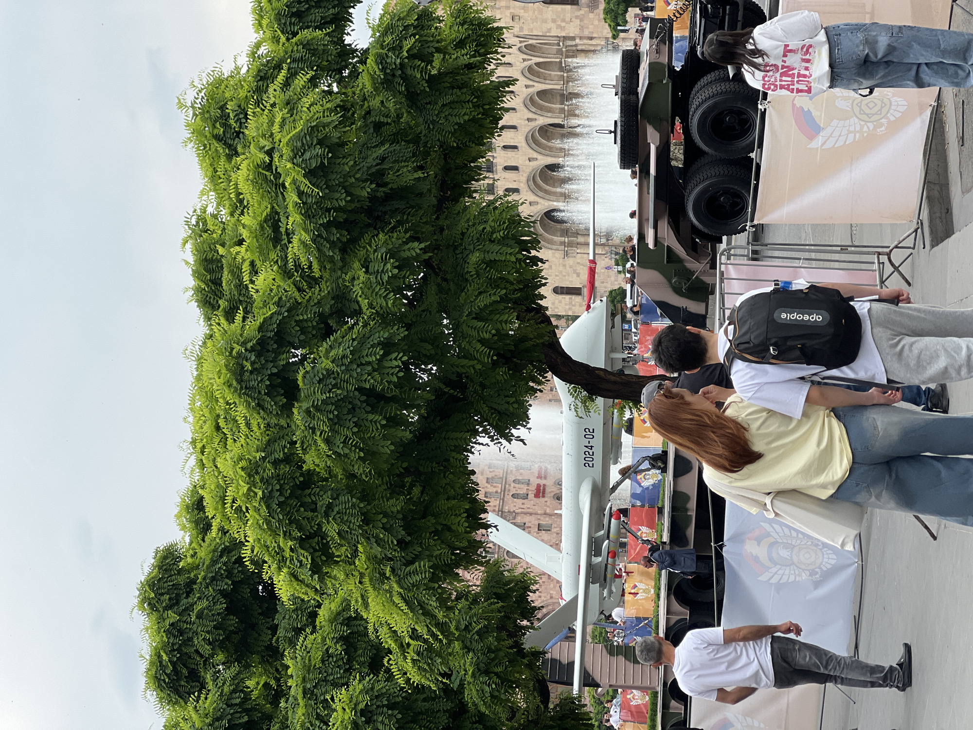 The image shows a public exhibition in Yerevan, Armenia. A large green tree dominates the left side of the frame. Behind the tree, there is a building with an arched facade and a fountain. An unmanned aerial vehicle is displayed near the tree, with the number "2024-02" visible on its body. To the right, there is a military truck with large tires, along with displays or banners with the flag or insignia of Armenia. Several people, including adults and children, are visible in the scene, some looking at the exhibits. The people are dressed casually and walking around the park and interacting with the display items.
