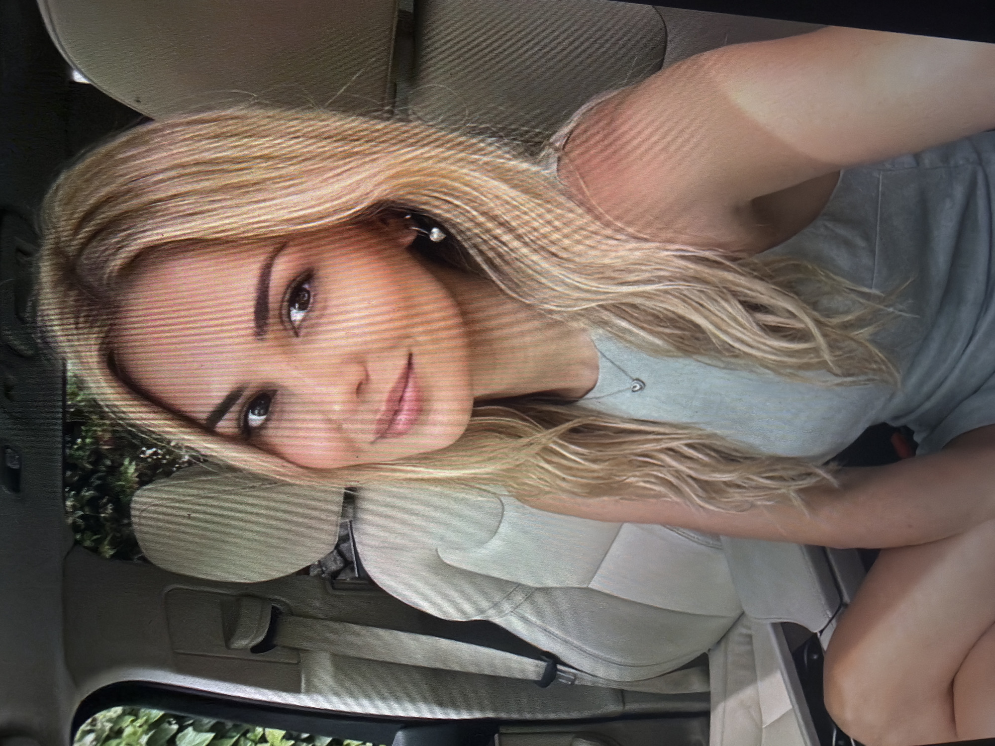 The image depicts a female individual positioned in the front passenger seat of a vehicle. The individual, a young adult female, has long, wavy blonde hair styled over her shoulders. She has dark eyebrows, dark eyes, and light-colored lipstick, with a slight smile directed towards the camera. She is wearing a light-colored, possibly light gray or pale blue, sleeveless top or dress. A small heart-shaped pendant necklace is visible on her chest, and she wears small hoop earrings, one of which appears to have a small, light-colored spherical drop.
The vehicle's interior features light beige or off-white upholstered seats. The passenger seat's headrest and a portion of the driver's seat, including its headrest and a dark-colored seatbelt, are visible. Through the vehicle's passenger side window, dense green foliage, indicative of trees or bushes, is discernible. The scene is illuminated by natural daylight.
There are no discernable landmarks, architectural features, or specific signage within the image that allow for a precise identification of a location within the city of Shumen, Bulgaria.