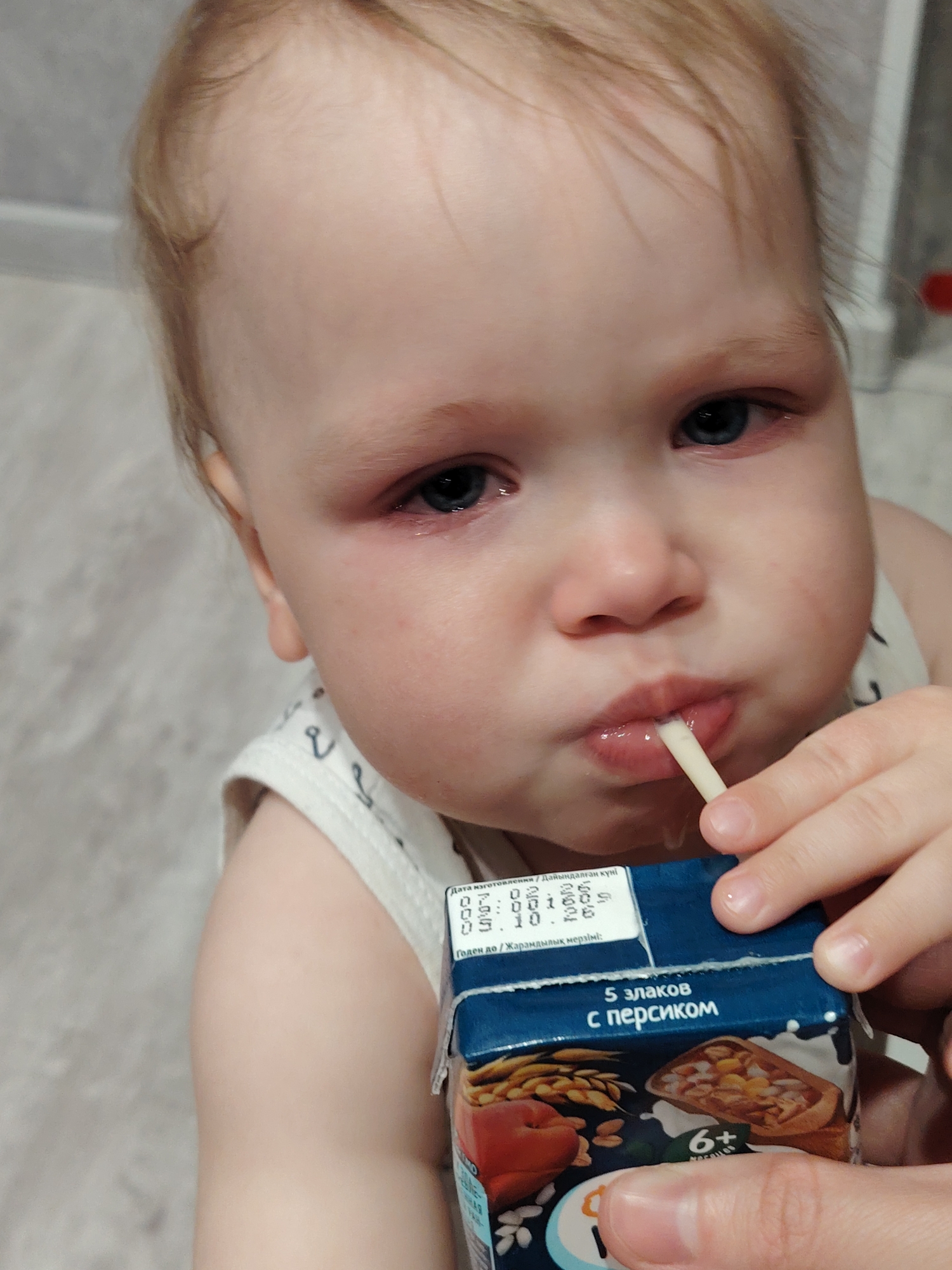 A close-up photograph features a young child with fair skin and light brown hair drinking from a small blue and white juice carton with a straw. The child's eyes appear watery, with redness around the eye area. The carton depicts a graphic of grains, a peach, and a cereal-like topping in a slice of bread or pie. Text on the carton, in Cyrillic script, indicates "5 злаков с персиком" ("5 cereals with peach") and "6+" possibly indicating the age range. A sticker on the carton displays dates, indicating "Дата изготовления" ("Date of manufacture") and "Годен до" ("Good until"). The background appears to be a neutral-toned wall. Given the Cyrillic text on the carton, it can be inferred the location is likely in a region that uses the Cyrillic alphabet, potentially Russia or another Slavic country. However, there is no confirmation of the exact location of Chaikovsky, Russia.