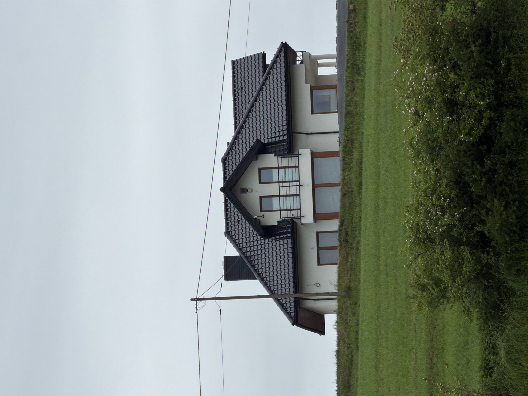 A detached, modern, two-story house with a black tiled roof and white walls stands on a slight incline in Męcina, Poland. The house features multiple windows, some of which are large and floor-to-ceiling, and a balcony with a black metal railing. A black chimney rises from the roof. The house is situated on a green hill, with a lush field of grass in the foreground. A cluster of bushes with small white flowers is visible in the lower right corner. A wooden utility pole with electrical wires running across it is positioned to the left of the house. The sky is overcast and gray.