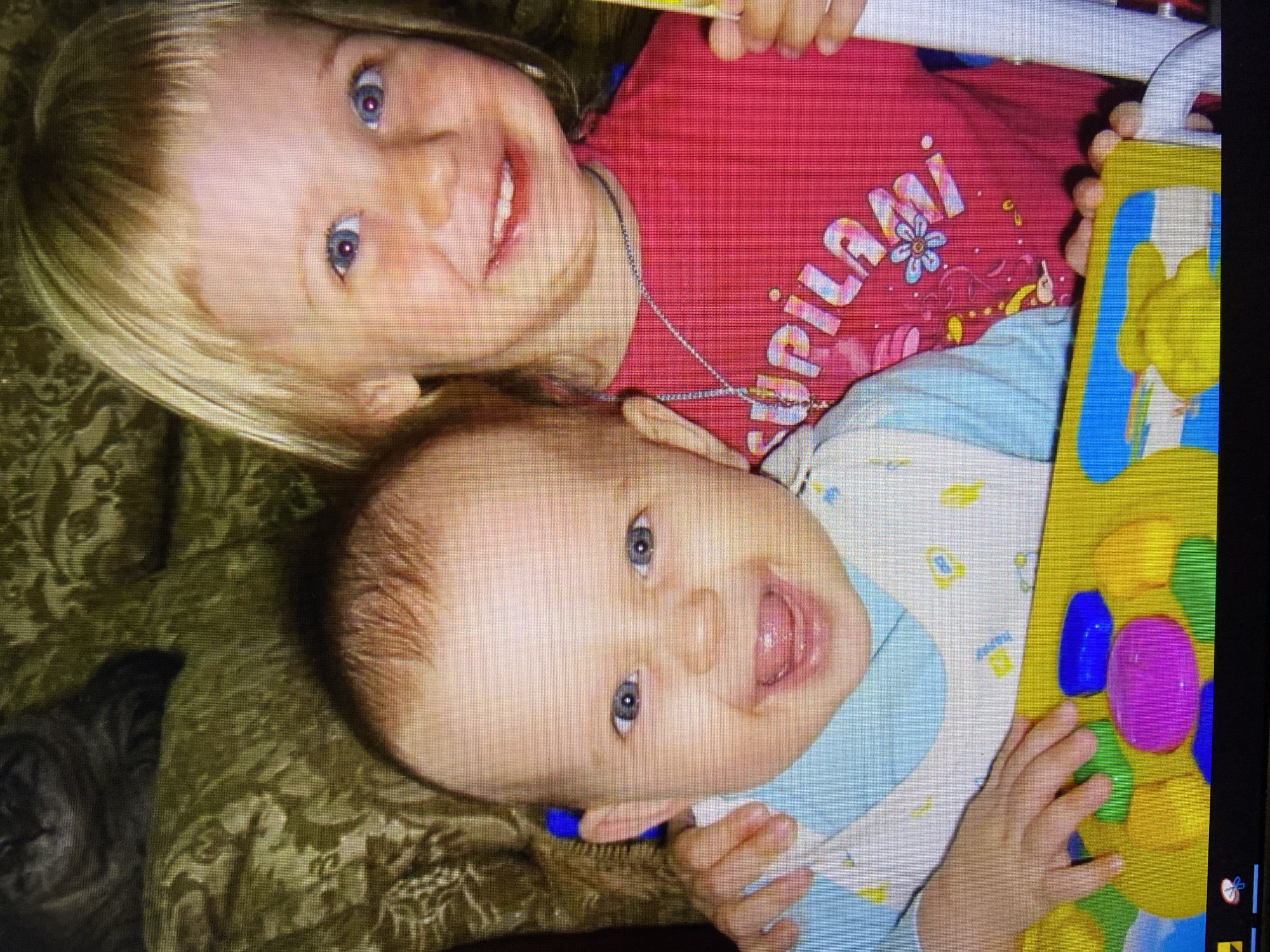 The image displays a close-up of two young children, likely siblings, posing for a photograph. The older child, a girl with blonde hair, smiles broadly at the camera, wearing a red t-shirt with a colorful graphic and text. Beside her, a younger child, a baby, also smiles, with their mouth open and tongue visible, wearing a light blue shirt with a white bib featuring colorful patterns. Both children have blue eyes. They are positioned in front of a textured green background, possibly a couch with a patterned fabric. A portion of what appears to be a toy, a bright yellow object with colorful buttons, is visible at the bottom of the frame, held by the children. The overall lighting suggests an indoor setting. The image quality is low resolution, with visible pixelation. No specific location in Stavropol, Russia, can be determined from the visual information provided.