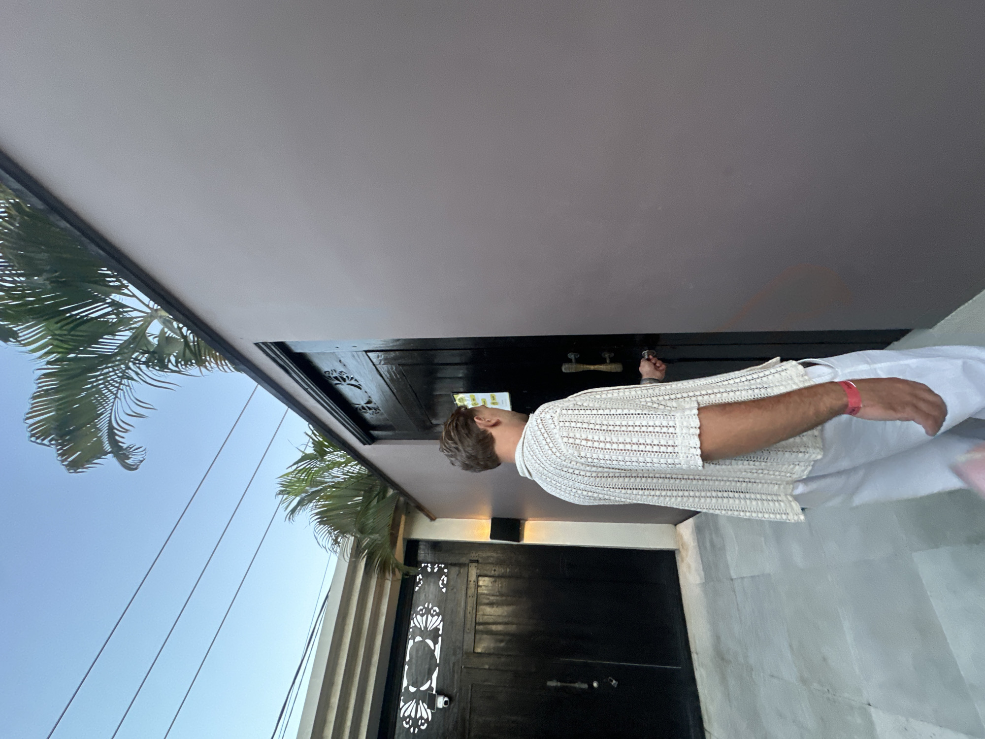 A man with short brown hair wearing a white crochet shirt and white pants is unlocking a black wooden door with decorative cutouts. He is standing on a light gray stone surface. Above the door is a gray awning. A palm tree is visible to the left, with a blue sky and power lines in the background. The location is likely North Kuta, Indonesia based on visual cues like the architectural style and the presence of palm trees.