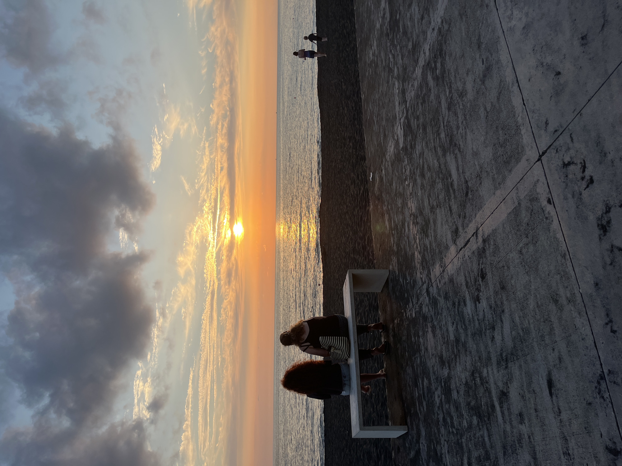 In Ureki, Georgia, two women sit on a concrete bench facing a dark sand beach and the Black Sea at sunset. The sky is a vibrant orange and yellow, with scattered clouds. The sun is low on the horizon, casting a shimmering reflection on the water. Two children and a woman are further down the beach. The foreground consists of a concrete paved area.