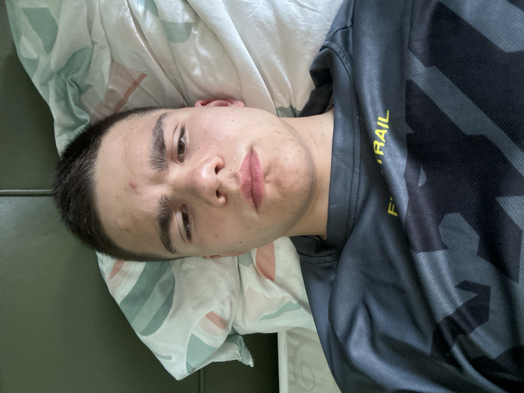 A young man is lying down, facing the camera. He is on a bed or similar soft surface with a patterned pillow under his head. He wears a dark, patterned athletic shirt with yellow lettering on the sleeve, partly obscured. The background includes a section of what appears to be dark furniture or a wall. The location is unknown.