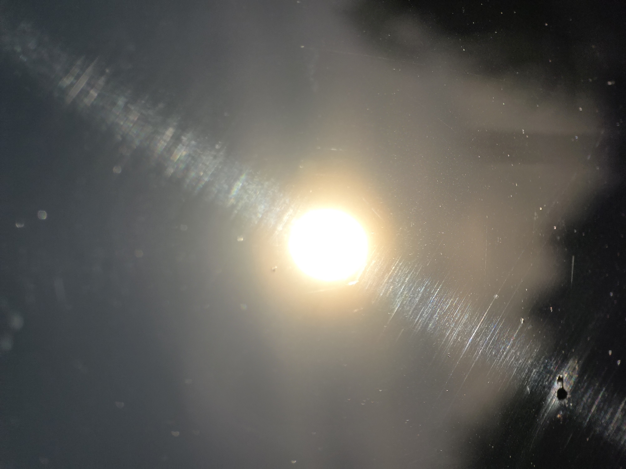 The image shows a bright light source, likely the sun or a strong artificial light, seen through a dirty, scratched surface, possibly glass. The surface is obscured by dirt, streaks, and what appear to be water droplets or imperfections. The overall scene has a hazy, unclear quality. The location and subject of the image in Chaikovsky, Russia are unknown.