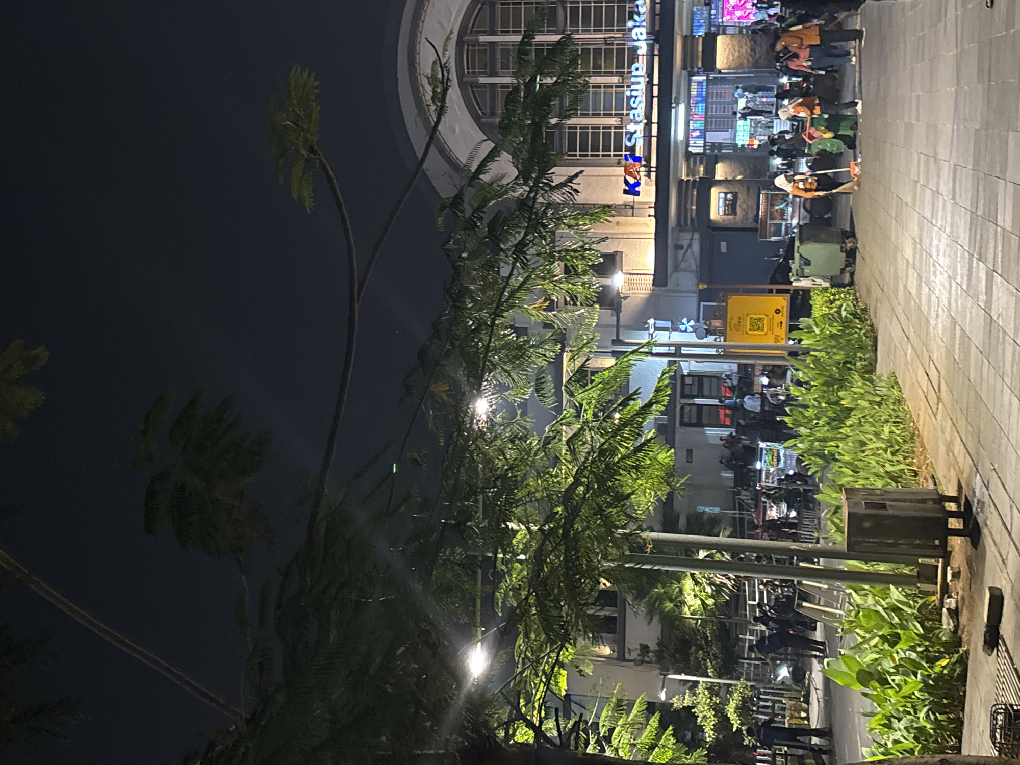 At night, a wide paved walkway leads towards the grand facade of the Jakarta Kota Station in West Jakarta, Indonesia. Lush green foliage lines the left side of the walkway, with tall trees whose branches hang over the path, illuminated by overhead streetlights. In the background, the station building features large arched windows and a prominent sign with blue neon lettering that reads "KA Stasiun Jakarta." People are visible on the station's plaza, some gathered around food stalls, others walking. On the walkway, a group of individuals in orange vests are sweeping and collecting trash near large green bins. The scene is well-lit, creating a vibrant atmosphere despite the darkness of the night.