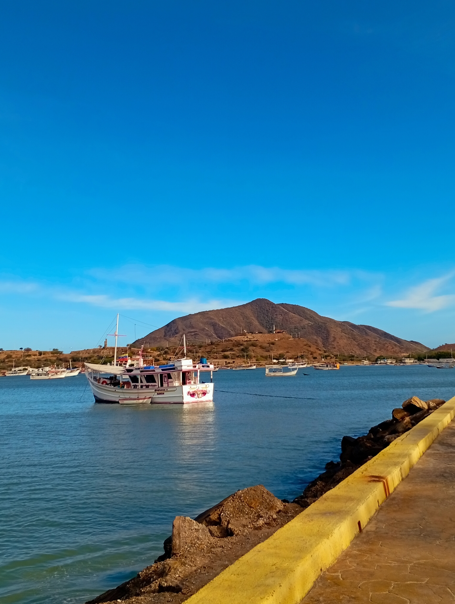 The image depicts a coastal scene in Juan Griego, Venezuela, under a clear blue sky with minimal white clouds. The foreground on the right features a concrete pier or seawall with a prominent bright yellow curb extending from the bottom right corner towards the mid-ground. Adjacent to the curb, dark brown rocks form a riprap descending into calm, blue-green water.
A white, multi-level boat with red trim and an enclosed cabin, marked with the name "MARICAR" on its stern, is anchored in the mid-ground. A smaller, white auxiliary boat is tethered to its side. Numerous other smaller boats, predominantly white, are visible anchored across the bay, particularly towards the distant shoreline on the left.
The background is dominated by a large, arid-looking brown mountain range with sparse vegetation, stretching horizontally across the horizon. Small structures, possibly buildings or communication towers, are discernible on its slopes and peak. No individuals, specific actions, or interactions are visible within the frame.