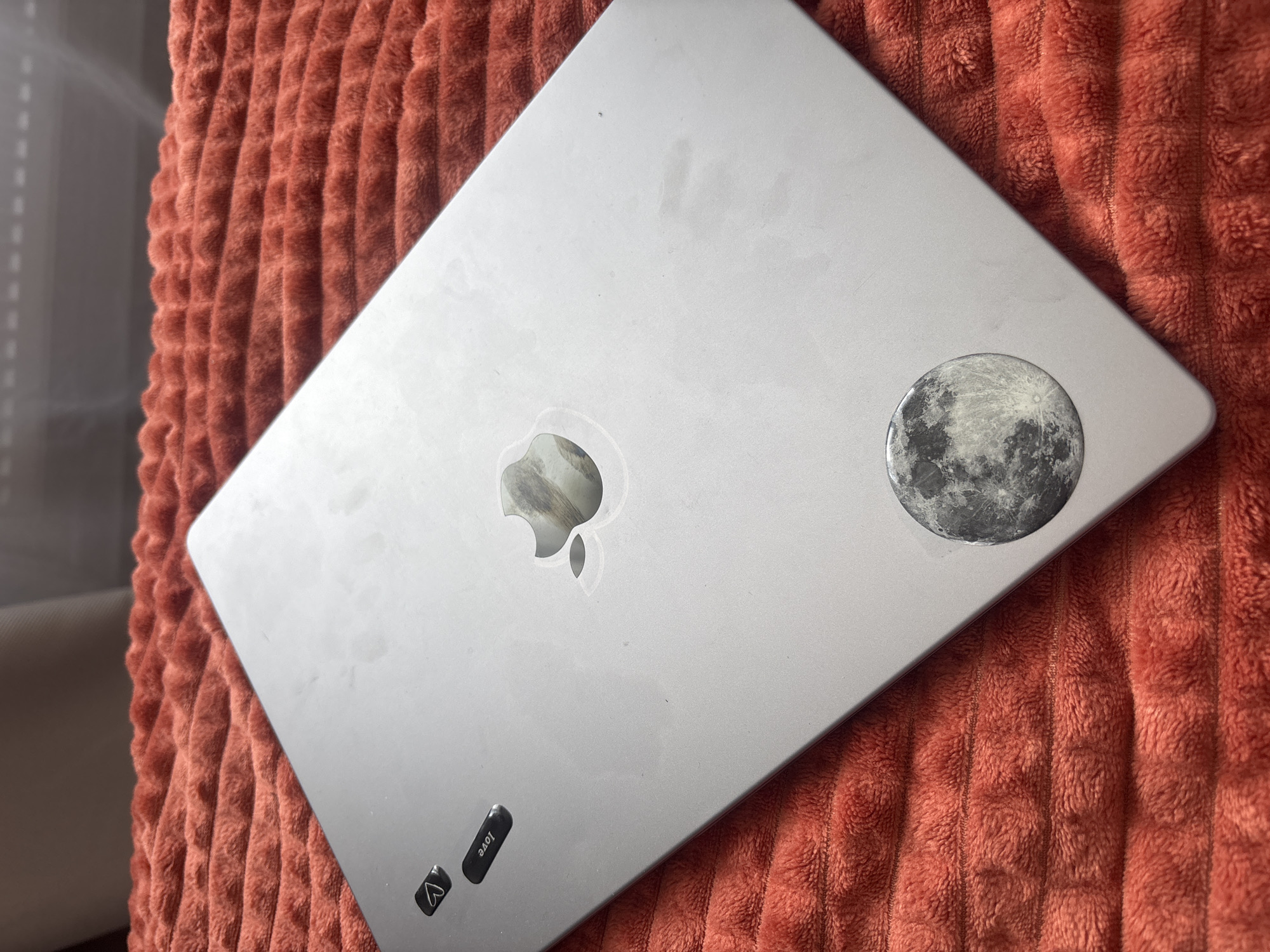 A slightly angled, overhead shot shows a closed, silver MacBook resting on a textured, orange, corduroy-like surface. The Apple logo is centrally located, and a round sticker depicting the moon is on the upper right corner. A small, dark sticker with the word "love" and a heart symbol is visible in the lower left corner of the laptop. The background consists of the textured orange surface, with a partial view of a light-colored upholstered object in the distant background. The city and specific location are unknown.