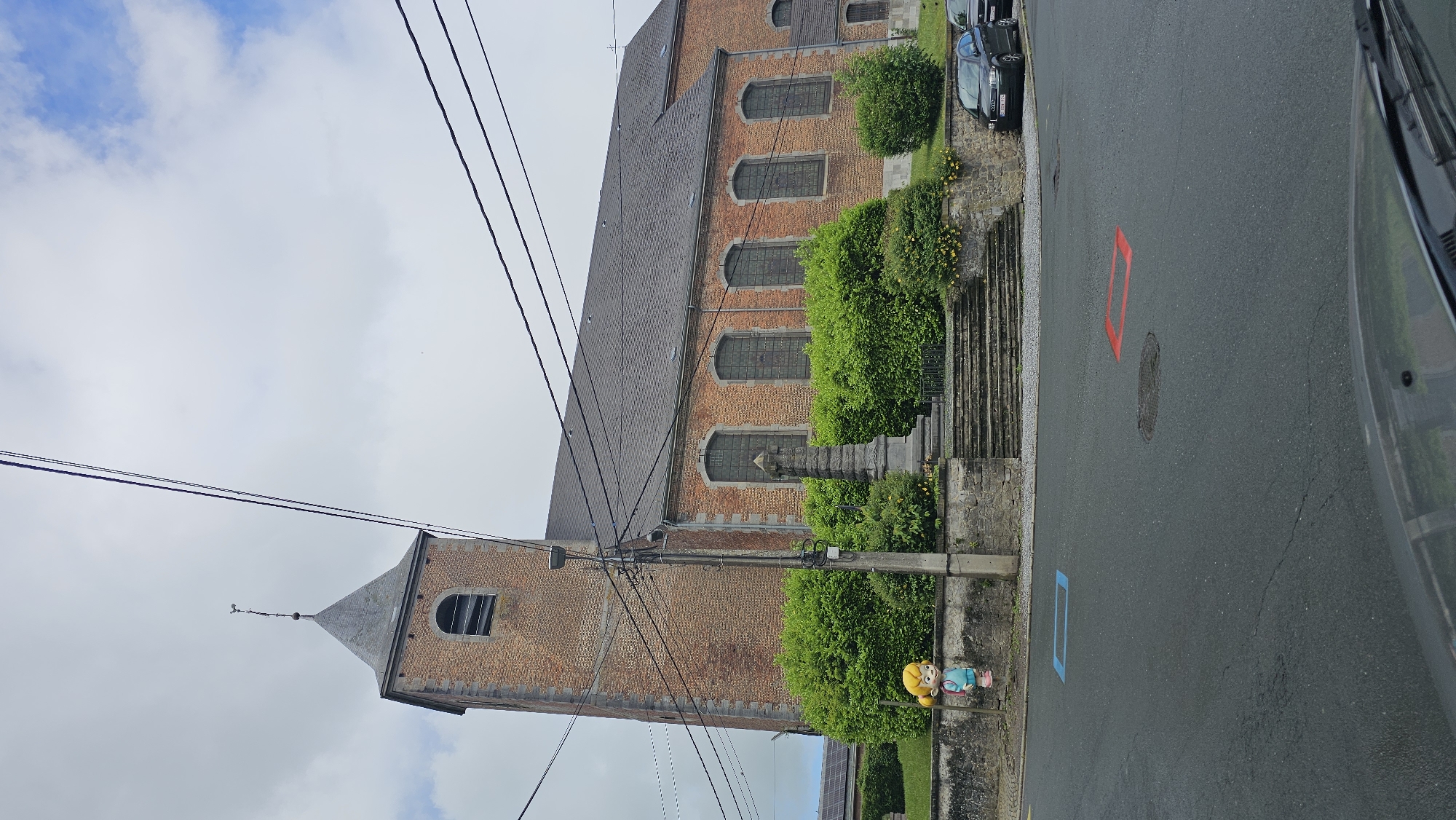 This daytime image, taken in Thuin, Belgium, presents a street scene dominated by a large, traditional brick building, likely a church or historical structure, on the left. The building features a dark shingle roof, several arched windows with dark panes, and a prominent steeple with a pointed roof and a single arched window.
In front of the building, a well-maintained garden with vibrant green bushes and some yellow-flowering plants borders a stone retaining wall. A set of stone steps leads from the street up to the building's level. A small, colorful figurine, depicting a cartoonish character with yellow hair, a blue top, and pink shoes, stands on the stone wall in the lower left.
The foreground consists of a paved road that appears damp, suggesting recent rain. Utility poles and numerous black overhead wires crisscross the frame, particularly in the mid-ground. On the right side of the road, several cars are parked, and the front corner of a vehicle, from which the photo was likely taken, is visible in the extreme right foreground.
Notable details on the road include a distinct red rectangular outline painted on the asphalt, with a blue rectangular outline visible further down the street. The sky is largely overcast, though patches of brighter cloud and some blue are discernible. The overall atmosphere is quiet and observational, capturing a typical street view in a European town. In the lower left background, beyond the garden, solar panels are partially visible.