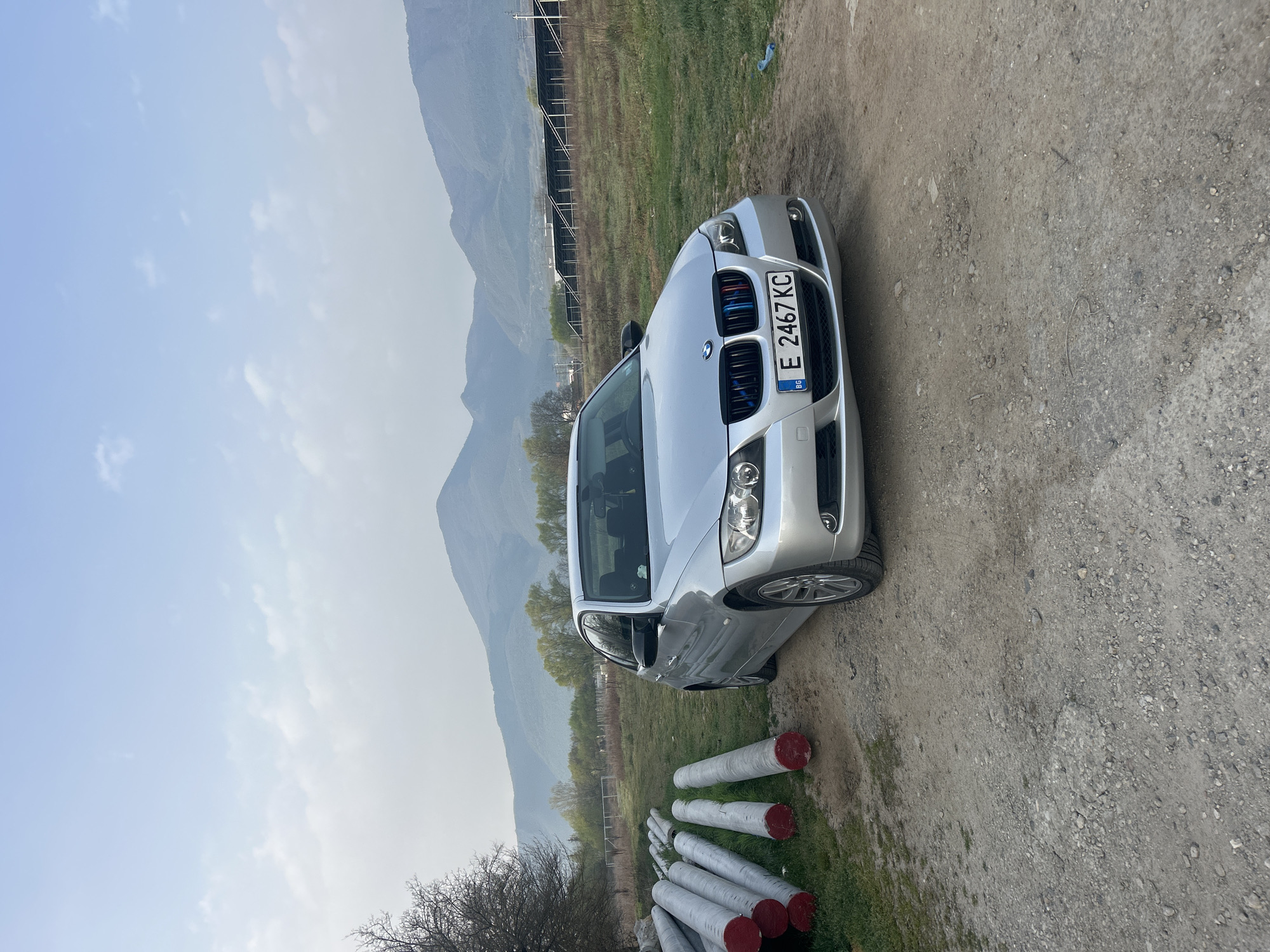 The image shows a silver BMW parked on a dirt road in an outdoor environment. The car's license plate reads "E 2467 KC". In the background, there is a mountainous landscape and a field with what appears to be agricultural infrastructure. In the foreground, there are several white cylindrical objects with red ends, possibly pipes or posts, lying on the ground near the car. The location is Unknown, Unknown.