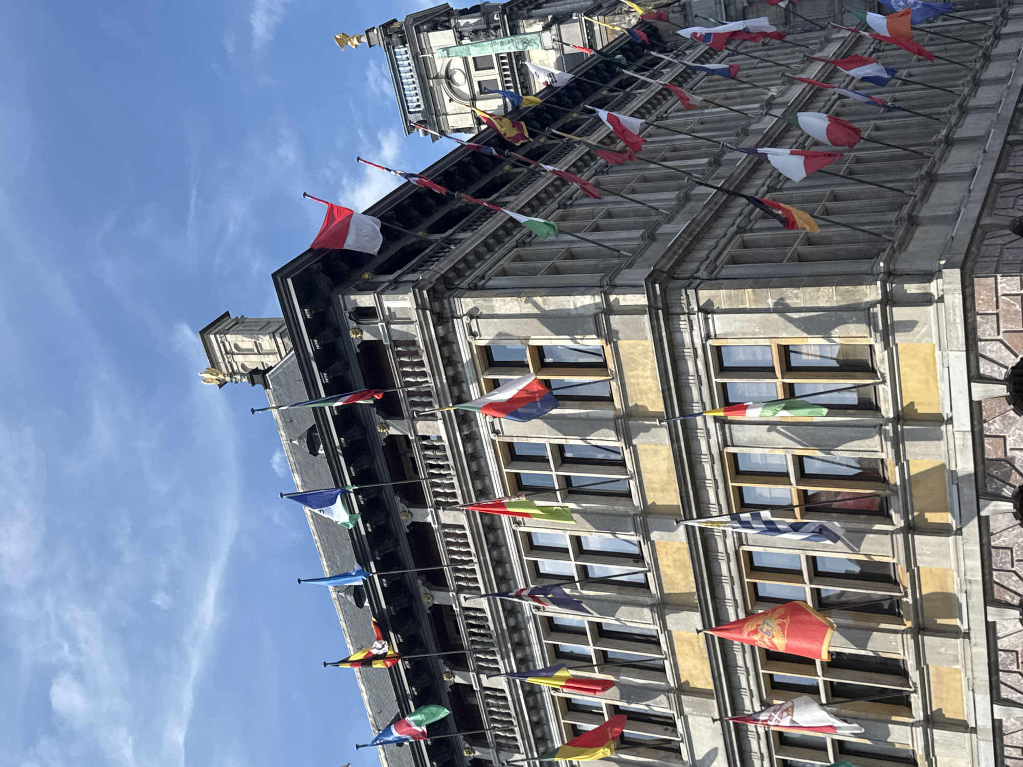 This is a close-up, eye-level, outdoor photograph of the Antwerp City Hall, located in the Grote Markt square of Antwerp, Belgium. The building is a Flemish Renaissance architectural masterpiece, adorned with numerous national and regional flags displayed on poles along its facade and upper levels. The sky above is a clear, bright blue with a few wispy white clouds. The flags, in various colors and designs, are fluttering in the breeze, creating a vibrant and patriotic atmosphere. The detailed stonework of the building, including its ornate windows and balconies, is clearly visible. No people are discernible in the image.