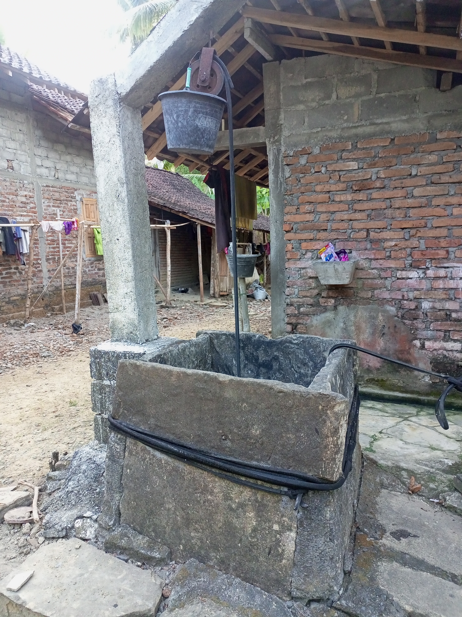 The image shows an outdoor scene in the city of Nanggulan, Indonesia. In the foreground, a concrete wellhead is visible, with a large stone rim. A black hose is wrapped around the wellhead. Behind the wellhead, a simple structure with a thatched roof and brick walls stands. A pulley system with a black bucket is suspended from an overhead beam, suggesting the well is in use. Clothing hangs on a clothesline to the left of the well, and several buildings with similar architectural styles are visible in the background. The ground is covered in sand and debris. The scene appears to be a residential area.