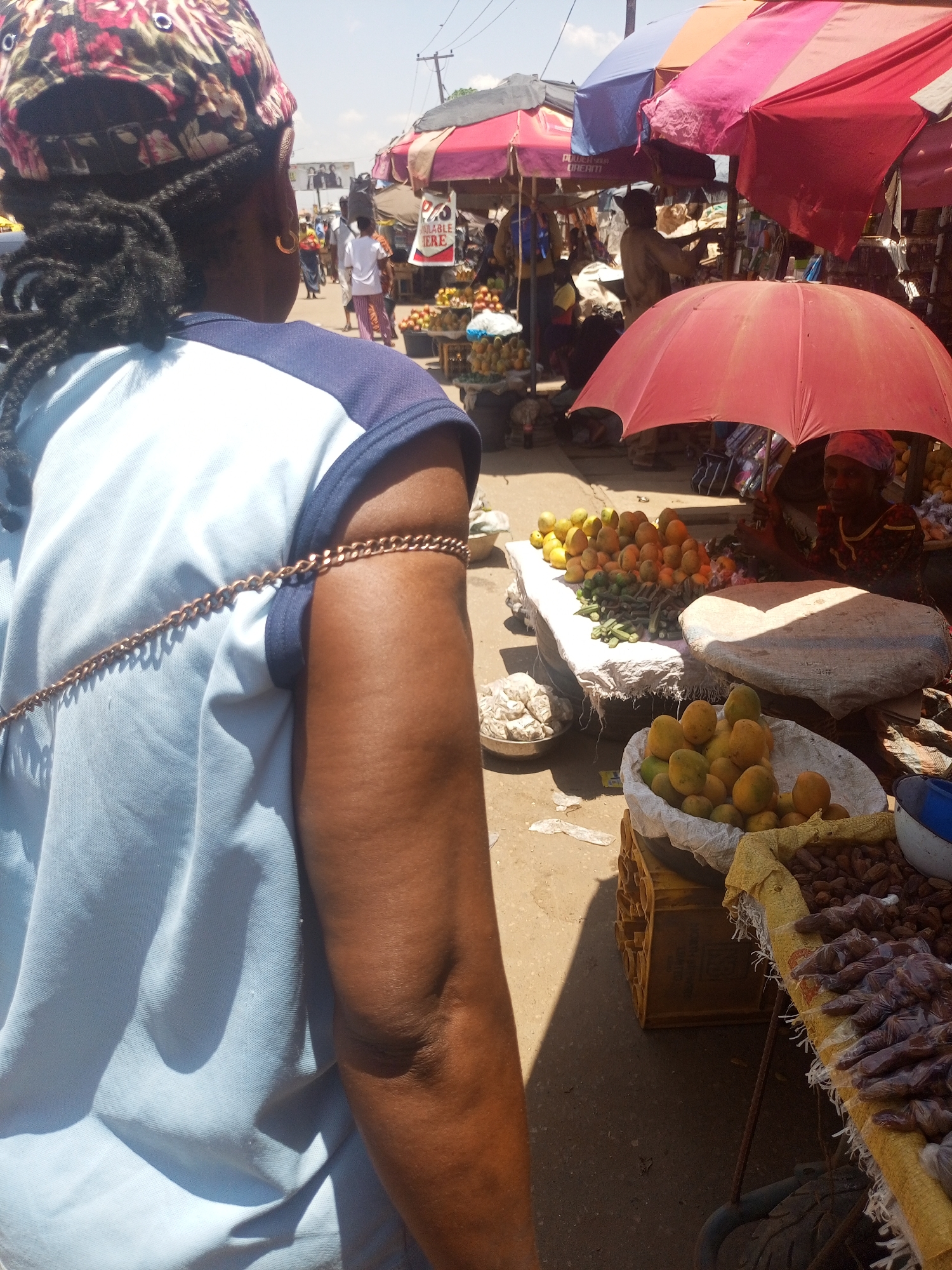 A medium, outdoor shot taken in Sabon Tasha, Nigeria, shows a person in the foreground with their back to the camera, wearing a light blue shirt with navy blue trim, a floral print hat, and a gold armlet. The foreground subject is facing toward a vibrant street market scene with multiple vendors under colorful umbrellas selling produce. A table with oranges, mangoes, and okra is visible, as well as dates wrapped in plastic. People are milling about in the background.