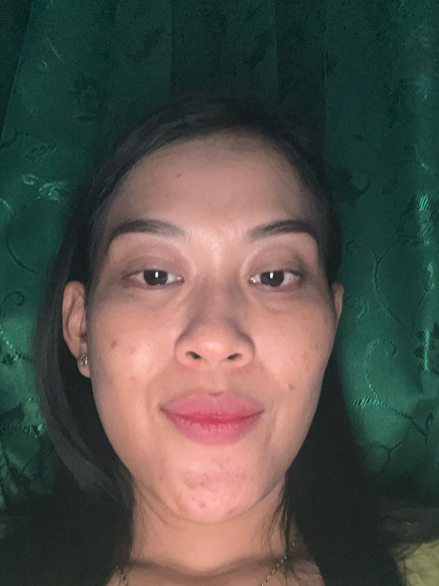 A close-up selfie of a woman with dark hair and dark eyes is captured against a dark green, textured background. Her skin tone is medium, with visible pores and some blemishes on her cheeks and chin. She is wearing light pink lipstick. A small gold hoop earring is visible in her left ear. She is wearing a necklace with a delicate chain. The lighting is from the front, casting soft shadows. The image does not provide any specific details or context that would allow for its location to be identified within Kecamatan Pati, Indonesia.