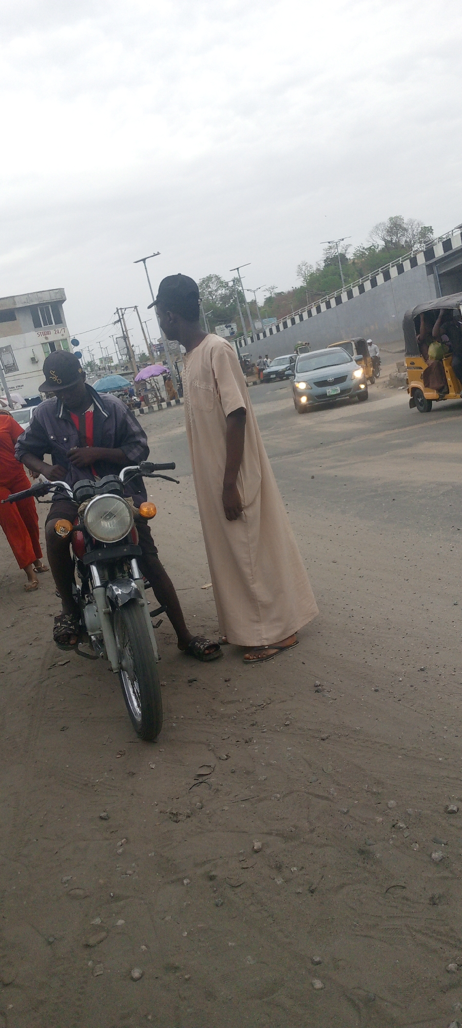 Two men are standing on a dirt road in Unknown, Unknown. One man is sitting on a motorcycle wearing a dark shirt and baseball cap. The other man is wearing a long, light-colored robe. There are cars, a tricycle, and buildings in the background.