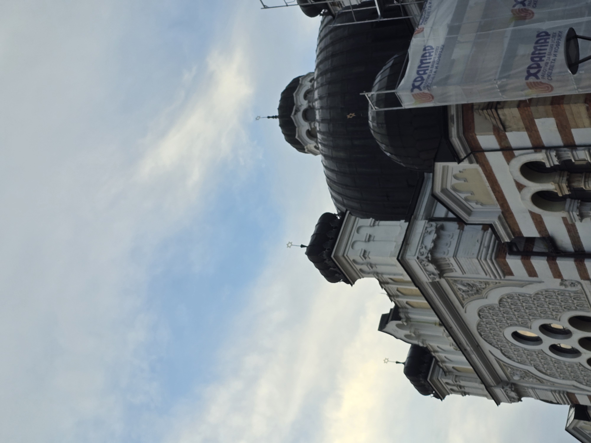 The image shows the Sofia Synagogue, a large Sephardi synagogue in Sofia, Bulgaria, set against a sky with white and blue clouds. The synagogue's architecture is visible, with intricate white and brown striped designs on the building and black domes topped with stars of David. A construction banner labeled "XPAMAD" hangs over the synagogue, suggesting that it is undergoing renovations. The angle of the photo is slightly tilted.