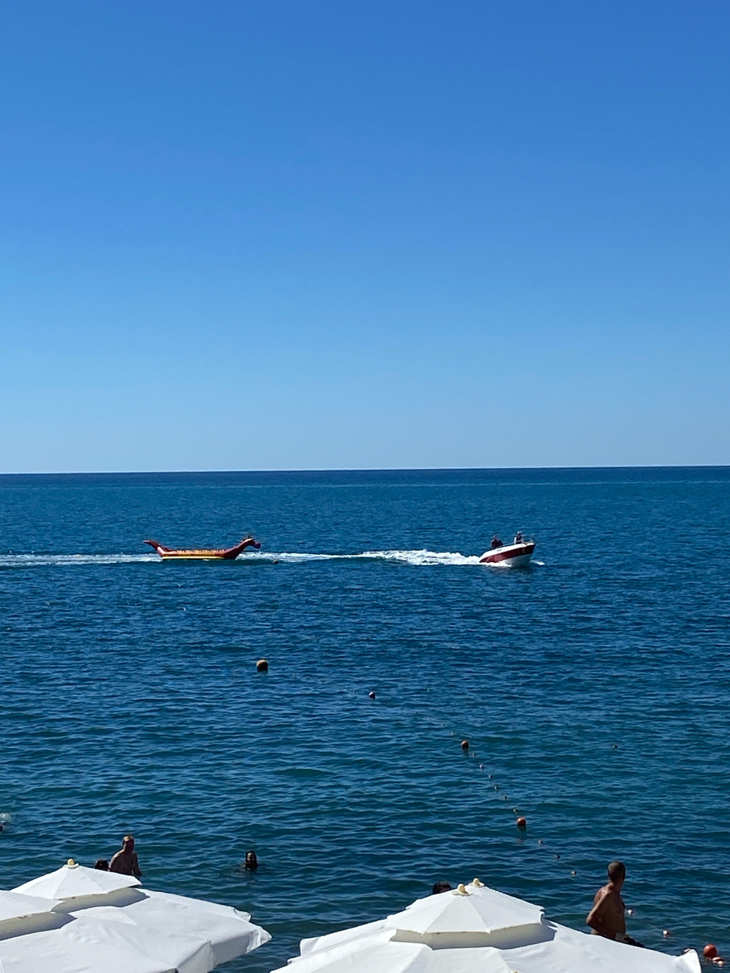 This is an eye-level, medium-shot photograph of the Black Sea coastline in Sochi, Russia, taken on a clear, sunny day. The sky is a vibrant, cloudless blue, and the sea is a deep, rich blue with moderate waves. In the distance, a speed boat with a white hull and red trim is moving from left to right, leaving a white wake. Two people are visible in the boat. Further to the left, a long, inflatable yellow and red banana boat, shaped like a dragon, is being towed across the water, also from left to right. There are a few buoys scattered on the water's surface. In the foreground, the tops of several white beach umbrellas are visible, suggesting a beach resort setting. A few people are in the water, with their heads and shoulders visible. One person is standing near the shore, and another appears to be swimming. The overall impression is a scene of leisure and recreation on a summer day.