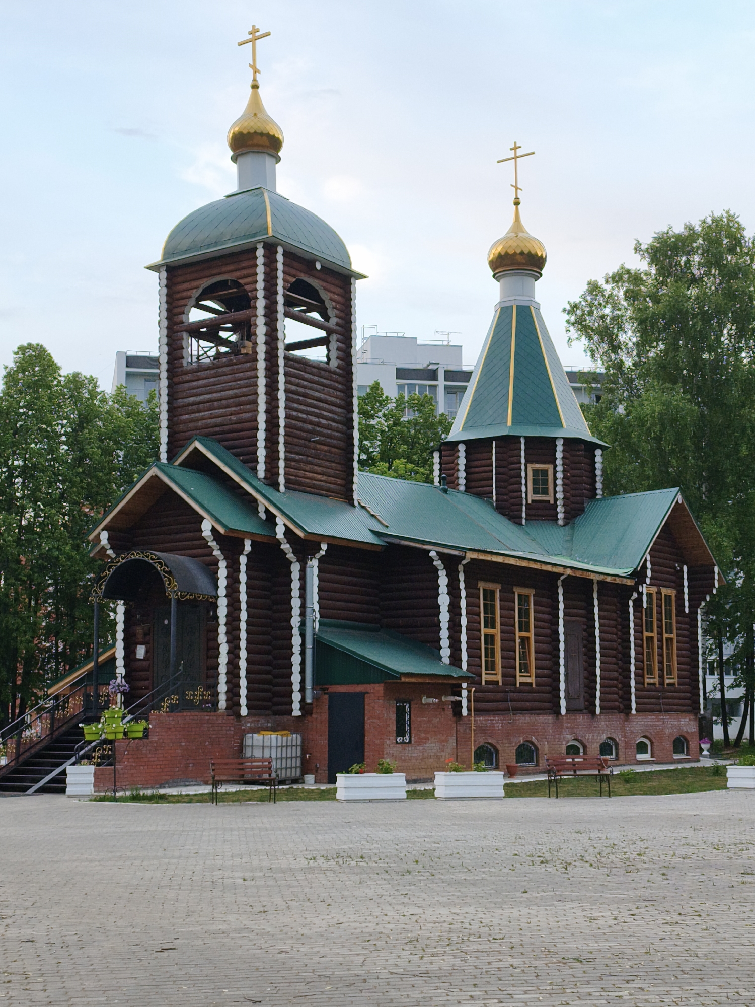 The image shows a wooden Orthodox church in Chaikovsky, Russia. The church is constructed of dark brown logs, accented with white trim. It features a green metal roof and two towers, each topped with a golden dome and a cross. The tower on the left is taller and includes an open belfry. A staircase leads up to the entrance on the left. A brick foundation is visible below the wooden structure. Benches and flower planters are placed around the perimeter of the church. The church is surrounded by trees. A modern building is visible in the background behind the trees. The foreground shows a paved area. The sky is overcast.