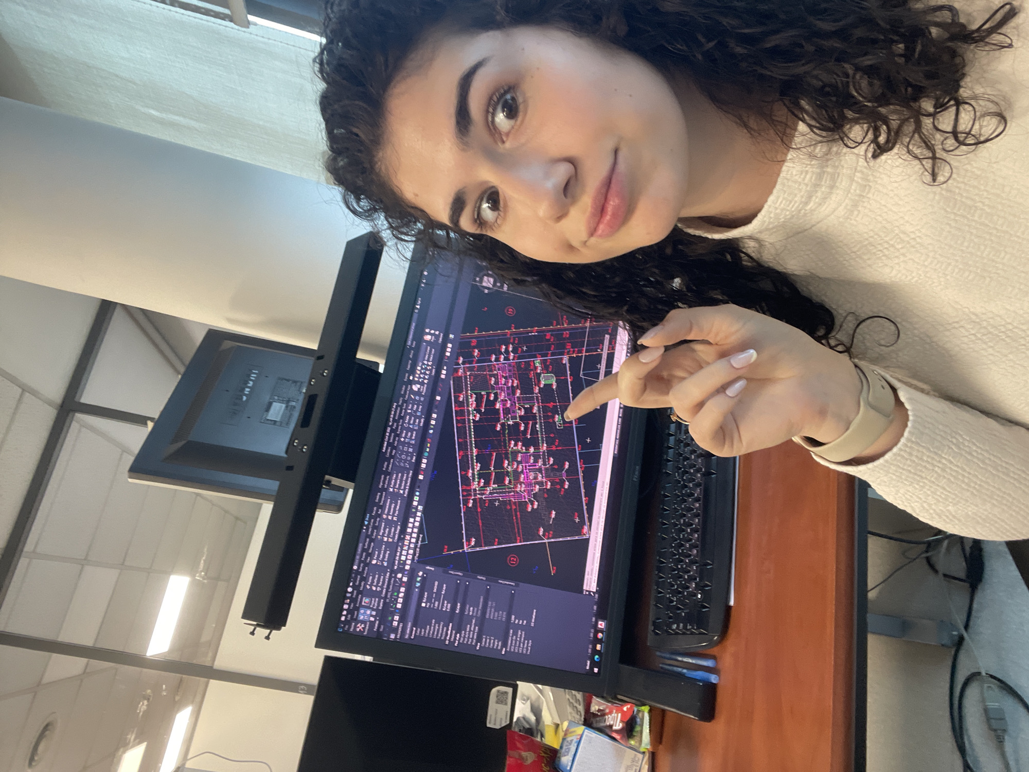 A young woman with dark, curly hair is shown taking a selfie in an office setting. She looks towards the camera with a subtle smile and an inquisitive expression. She is wearing a light-colored, textured sweater and has light-colored nail polish on her manicured nails. A light-colored watch or band is visible on her left wrist.
The woman is seated at a wooden desk with two computer monitors. Her right index finger points towards the main monitor, which displays what appears to be a complex engineering or architectural design within CAD (Computer-Aided Design) software. The screen shows intricate diagrams, lines, shapes, and small, unreadable text, indicative of a detailed layout or schematic. A black keyboard is placed next to this monitor. To the left, a second, dark monitor is visible, mounted on a stand.
The background reveals a typical office environment with white ceiling tiles, overhead fluorescent lighting panels, and office partitions. Below the main monitor, there is some clutter on the desk, including boxes of snacks, with the brand "Topps" clearly visible on one, along with various cables and a QR code sticker. The scene depicts a professional workspace, likely during daytime. The location hint indicates Sofia, Bulgaria.