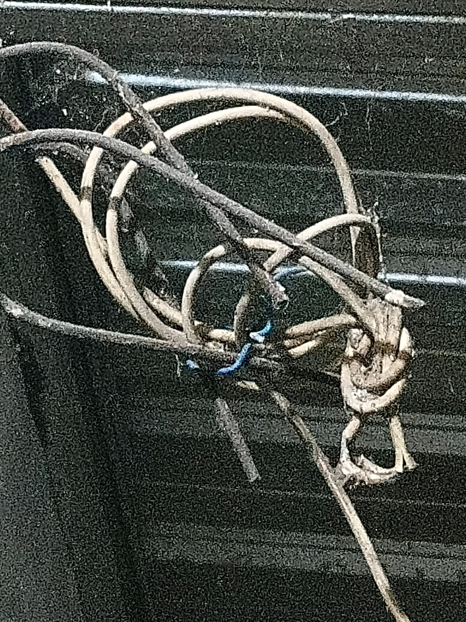 The image displays a close-up, overhead view of a tangled mess of wires. The wires are a mix of dark gray and a lighter tan color, with a few strands of blue wire visible. The wires appear to be old and frayed, with some of them knotted together. The background is a dark, textured surface with faint horizontal lines. There is no indication of location in the city of Cikal, Indonesia, or any people, actions, or interactions. The image is purely a detailed depiction of the wires.