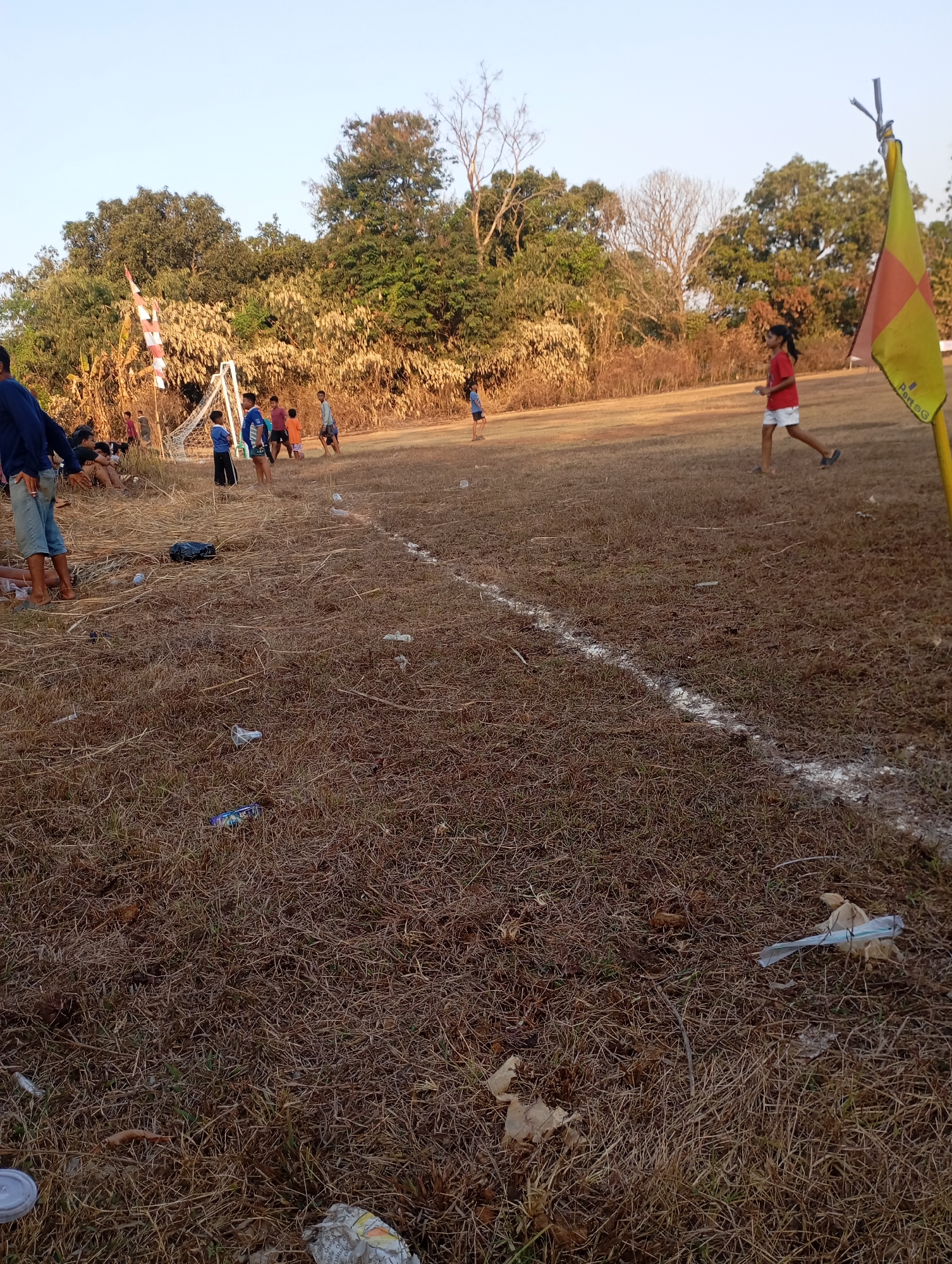 A group of children are playing soccer on a dry, grassy field in Kecamatan Haurgeulis, Indonesia. The field is uneven and littered with dry grass and some trash, including plastic bottles and bags. A soccer goal is visible in the background, along with a yellow and orange corner flag. The sky is clear and the sun is shining. Several children are actively playing soccer, while others are sitting on the sidelines, watching. Some adults are also present, observing the game. The scene appears to be a casual outdoor activity.
