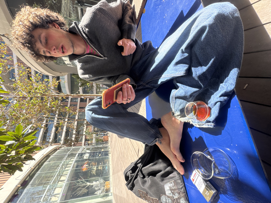 An individual is seated barefoot on a prominent blue outdoor surface, positioned adjacent to wooden decking, under direct sunlight. The person, appearing male with voluminous brown curly hair, is dressed in a charcoal grey quarter-zip sweater layered over a pink and white striped undershirt, and blue denim jeans. They hold an orange-cased smartphone with a visible gold card in their left hand. A silver hoop earring adorns their left ear, and their mouth is slightly agape. To their right, on the blue surface, rests a small, clear glass containing a reddish-brown liquid. To their left, near the bare left foot, a gold-colored cigarette pack (featuring Hebrew text) and a clear glass ashtray are present, alongside a black fabric bag with embroidered or printed details. The background comprises modern, multi-story glass-fronted commercial or residential buildings and trees with sparse foliage, consistent with the Sarona Market complex in Tel-Aviv, Israel.