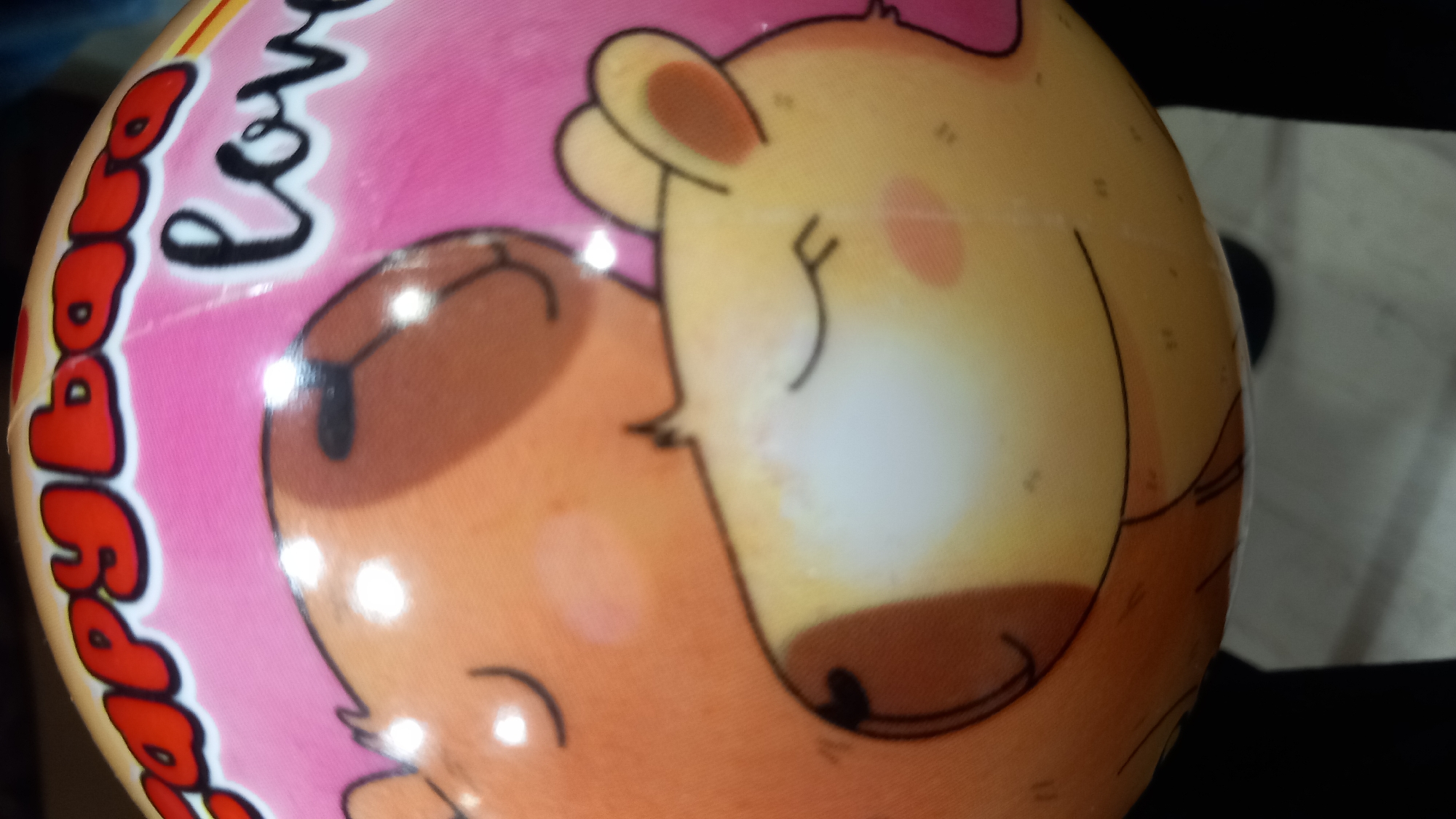 The image shows a close-up of a round object, likely a ball or a toy, decorated with cartoon capybaras and text. The capybaras are depicted in a loving embrace, with closed eyes and rosy cheeks. The text above them reads "Capybara love" in red, stylized lettering and cursive. The object is predominantly pink and yellow, with details in brown and black. There are no discernible elements in the image that suggest a specific location within the city of Kos, Greece.