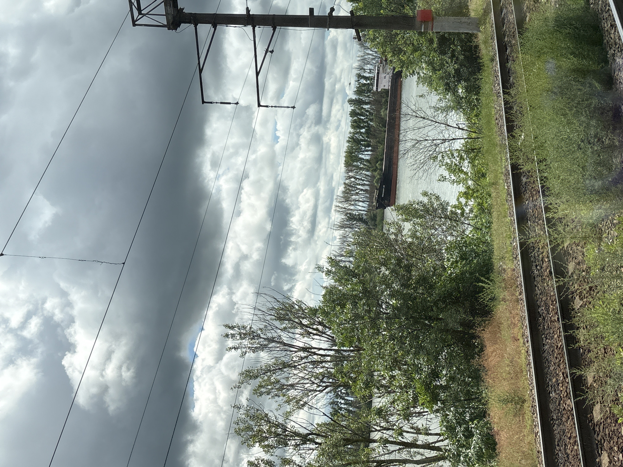 The image depicts a railway track running parallel to a body of water, likely the Don River, near Rostov-on-Don, Russia. A barge is visible on the water, positioned between a line of trees. Overhead electrical wires span the scene. The sky is overcast with dark, heavy clouds. Vegetation, including trees and ground cover, borders the railway track. The scene appears to be taken from a moving train.