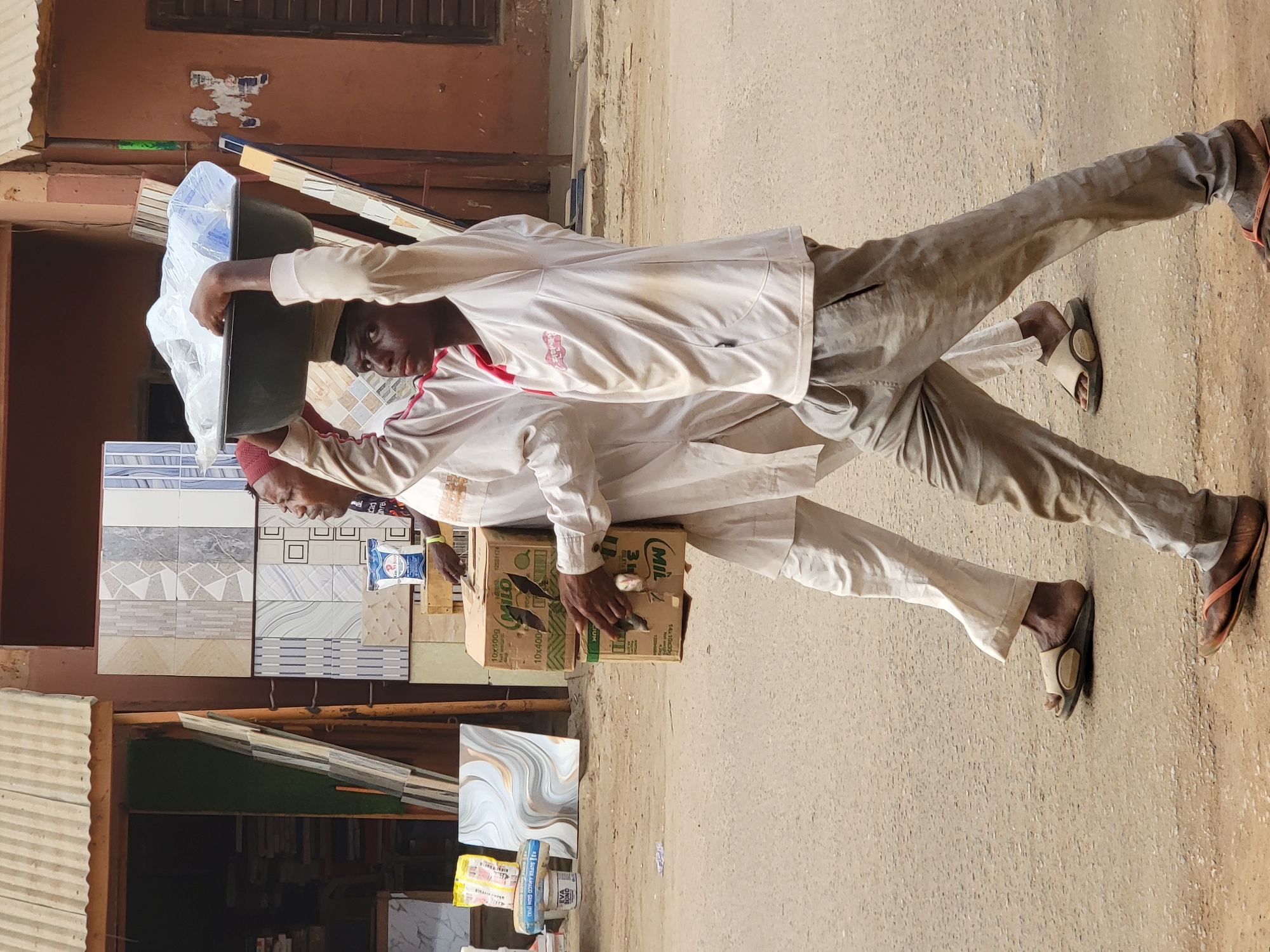 The image captures two men walking outdoors in what appears to be a street scene in Maiburiji, Nigeria, during bright daylight.
The man in the foreground is seen from the chest up, carrying a dark-colored, shallow basin on his head. The basin is covered with a clear plastic bag. He wears a light-colored, possibly white or off-white, short-sleeved shirt with red trim or accents, and light beige or brown trousers. His feet are visible, wearing brown flip-flops with a light-colored strap. He has dark skin and is looking slightly towards the viewer's left.
Behind him, partially obscured, is a second man wearing a white, long-sleeved garment, possibly a traditional jalabiya or agbada, and a red cap. He is carrying a large cardboard box in his arms. The box has the brand name "MILO" clearly visible, along with text indicating "3 IN 1" and what appears to be "10x400g" and "10x105g" on its sides. This man is also wearing flip-flops, light-colored with dark soles.
The background features a building with an orange-brown, somewhat textured wall. Below this, an array of patterned ceramic tiles are displayed vertically, suggesting a tile or building materials shop. A corrugated metal sheet is visible on the left side of the frame, possibly part of a stall or structure. The ground is a light-colored, paved surface, indicative of a street or pathway.
The men appear to be in the process of transporting goods, with one balancing a basin on his head and the other carrying a box. The bright, clear lighting suggests it is daytime, likely mid-morning or afternoon, with no strong shadows indicating early morning or late evening. The overall scene depicts daily life and commerce in a West African setting.