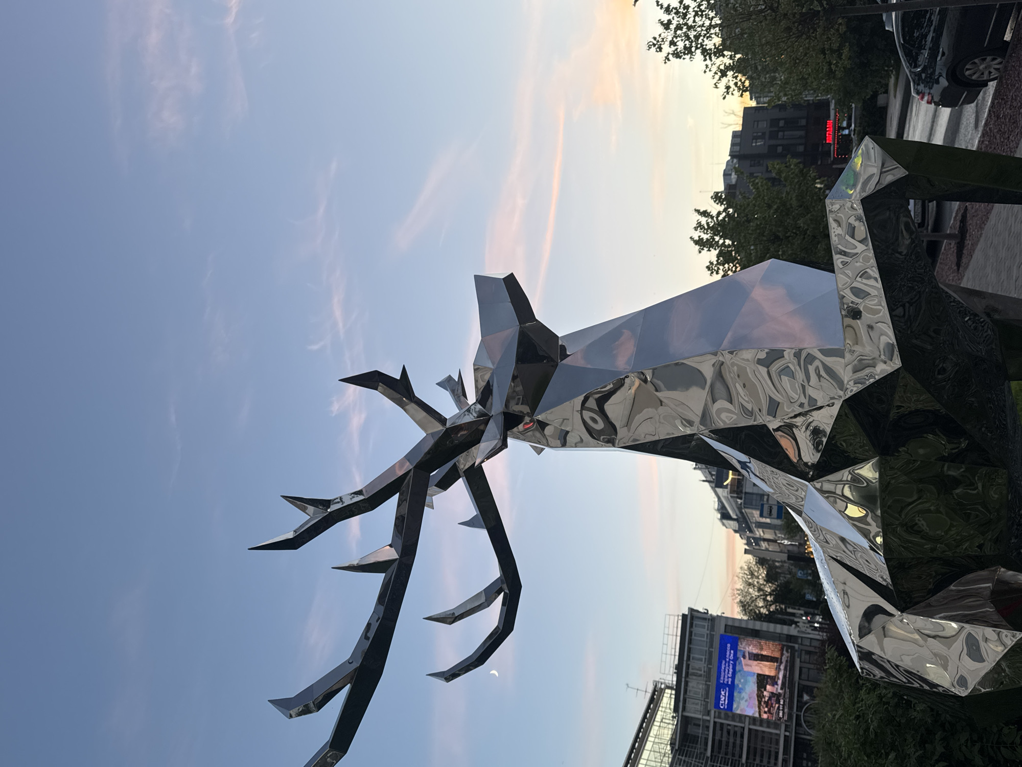 A large, faceted, chrome sculpture of a deer stands in a city setting against a twilight sky. The sculpture is geometric in design, with sharp angles and flat surfaces that reflect the surrounding environment. The deer sculpture is the dominant subject, positioned in the foreground and taking up a significant portion of the frame.
In the background, buildings and trees are visible, suggesting an urban location. A digital billboard displaying an advertisement is mounted on one of the buildings. The sky displays a gradient of colors, with hues of blue, pink, and orange, indicative of sunset or sunrise. A crescent moon is visible in the upper left quadrant of the sky. The scene appears to be a public space, possibly a park or a plaza, with a sidewalk and a street visible.
The location is the city of Nizhny Novgorod, Russia. Specifically, this sculpture is located on Bolshaya Pokrovskaya Street, a prominent pedestrian street in the city center. The sculpture is a modern art installation, often referred to as "Deer" or "Stag," created by artist Mikhail Shemyakin. It is a popular landmark and photo opportunity for tourists and locals alike.