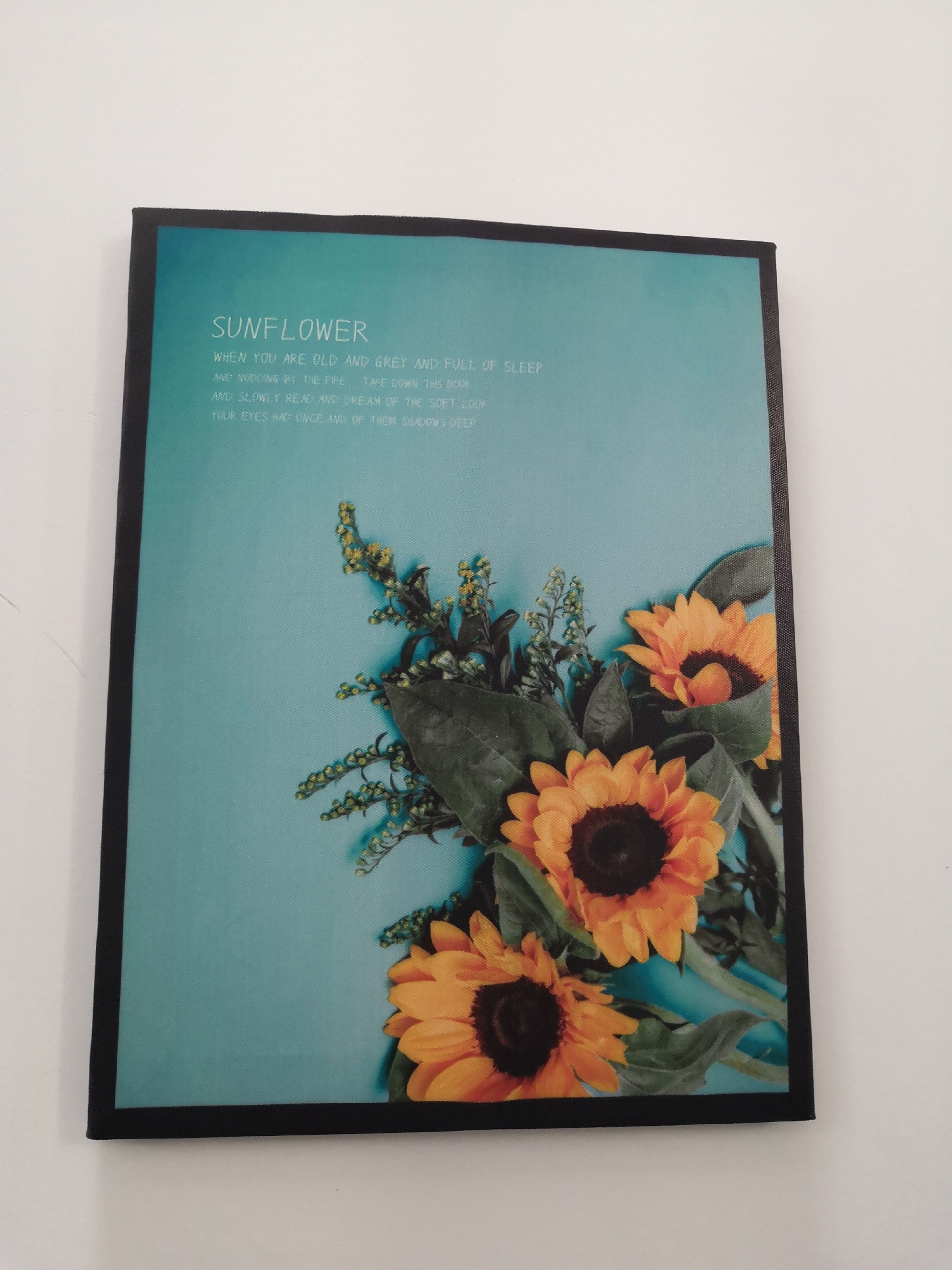 A framed picture is mounted on a white wall. The picture has a turquoise background with an image of sunflowers in the lower right corner. Written in white letters in the upper left corner are the words "SUNFLOWER" followed by a short poem. The frame around the picture is black.