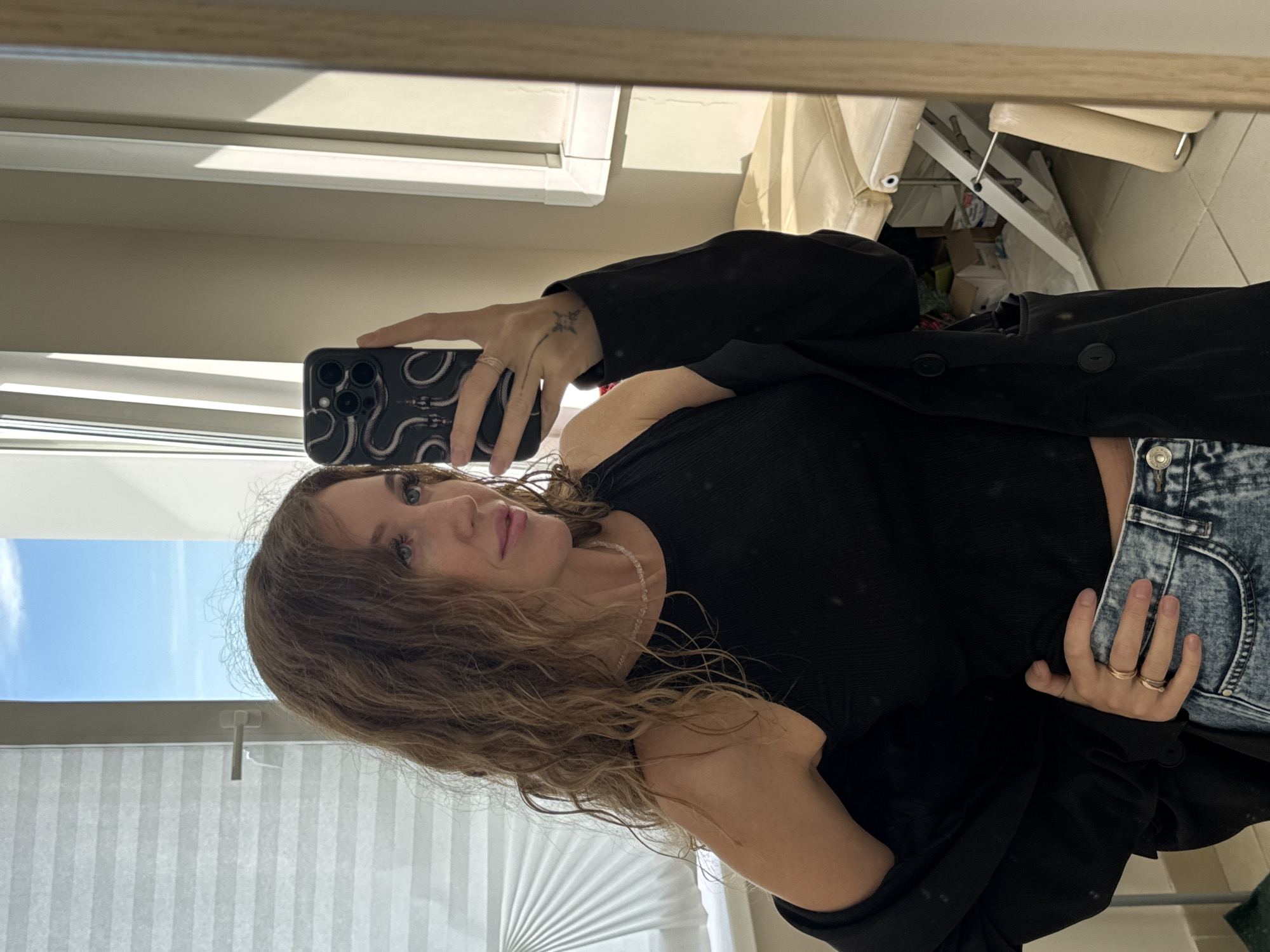 A woman is taking a mirror selfie in what appears to be a room with natural light. She is wearing a black tank top, a black blazer draped over her shoulders, and distressed blue jeans. She has curly brown hair and is wearing rings on her fingers and a necklace. She is holding a black smartphone with a snake-patterned case. In the background, a window with blinds is visible, and the room contains some furniture that resembles medical equipment, possibly in an office or studio setting. There is a mirror reflecting the scene. The location is specified as Chaikovsky, Russia.