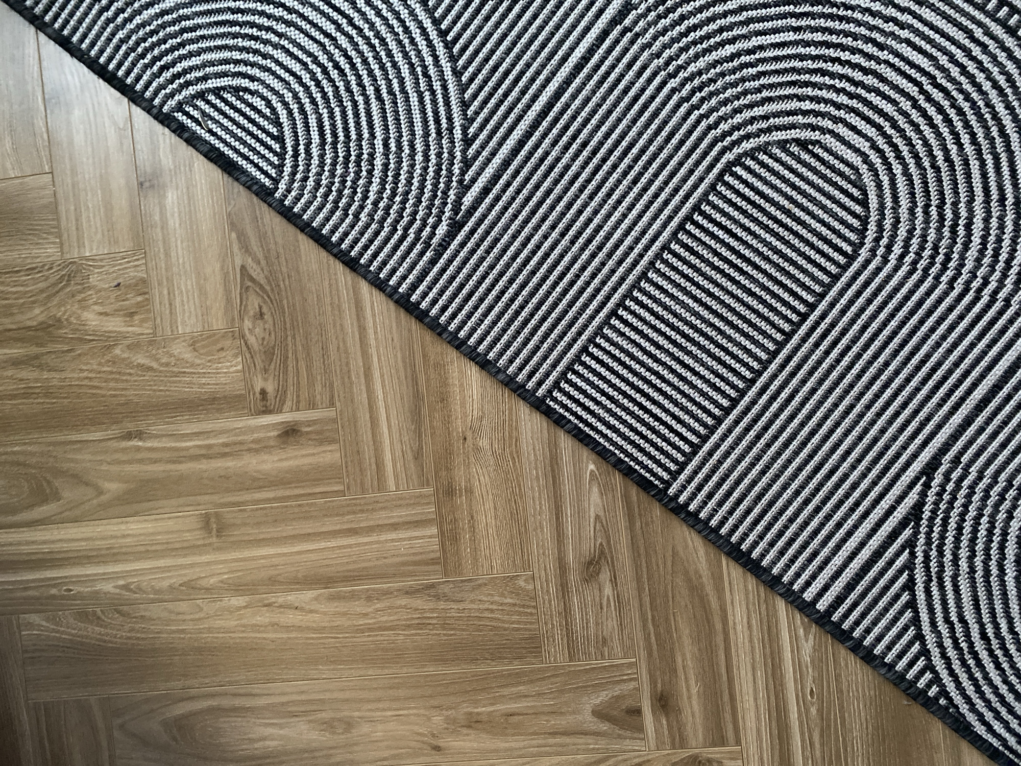 The image shows a section of a room with wooden flooring and a patterned rug. The flooring is laid in a herringbone pattern and appears to be made of wood or a wood-like material. The rug is positioned diagonally across the image, occupying the top right portion. The rug has a geometric pattern of repeating semi-circular shapes and parallel lines in alternating black and white stripes. There are no people visible in the image. The location is not discernible from the image alone.