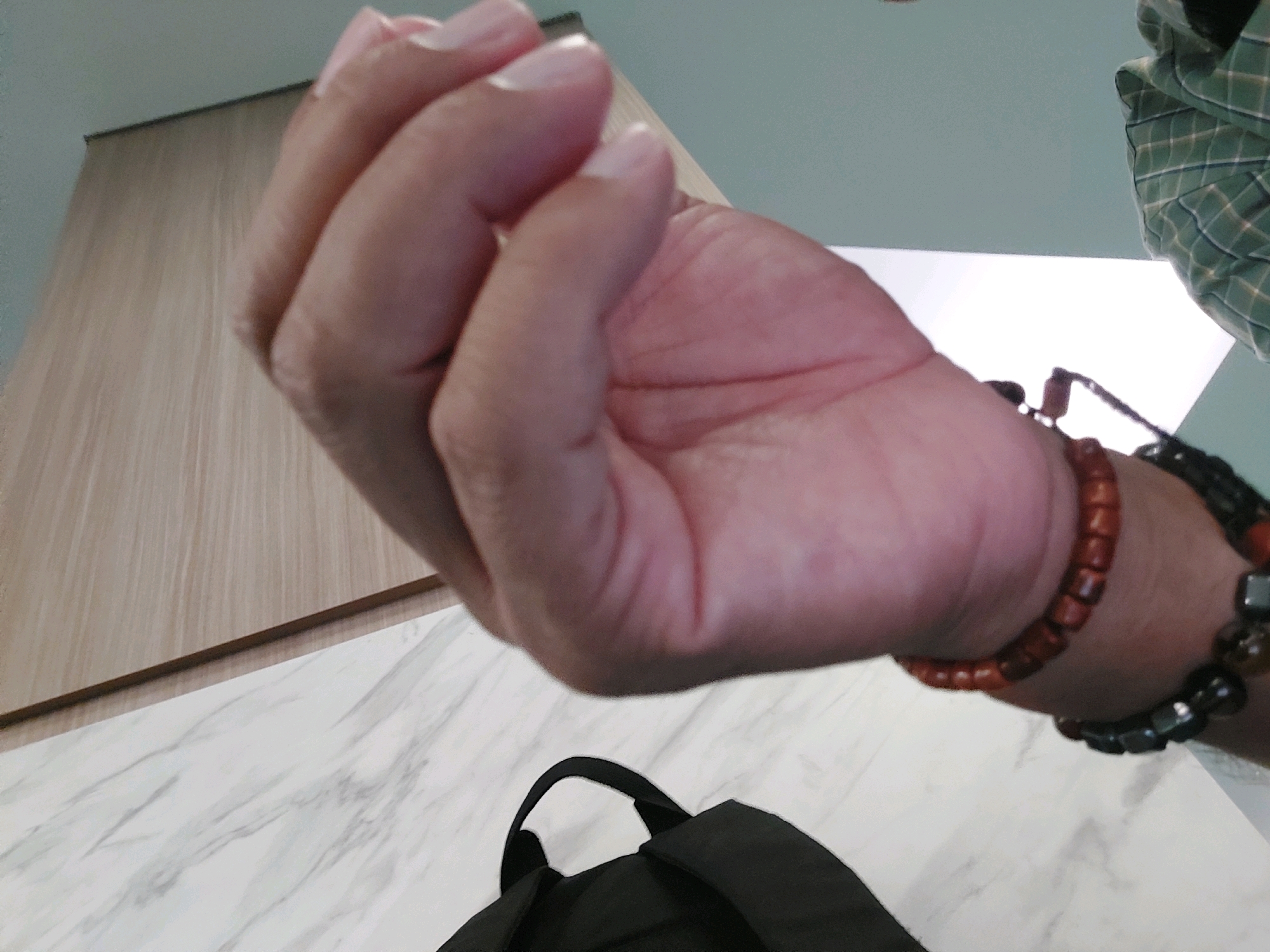 The image is an over-the-shoulder shot of a person's hand and wrist, viewed from a low angle. The hand is raised, with the palm facing downwards and slightly towards the viewer. The person is wearing bracelets on their wrist. In the background, there is a light-colored marble-patterned surface, a light wood-paneled wall, and a light blue ceiling. A black backpack is partially visible at the bottom of the frame. The image does not contain any elements that would allow for the determination of a specific city or country.