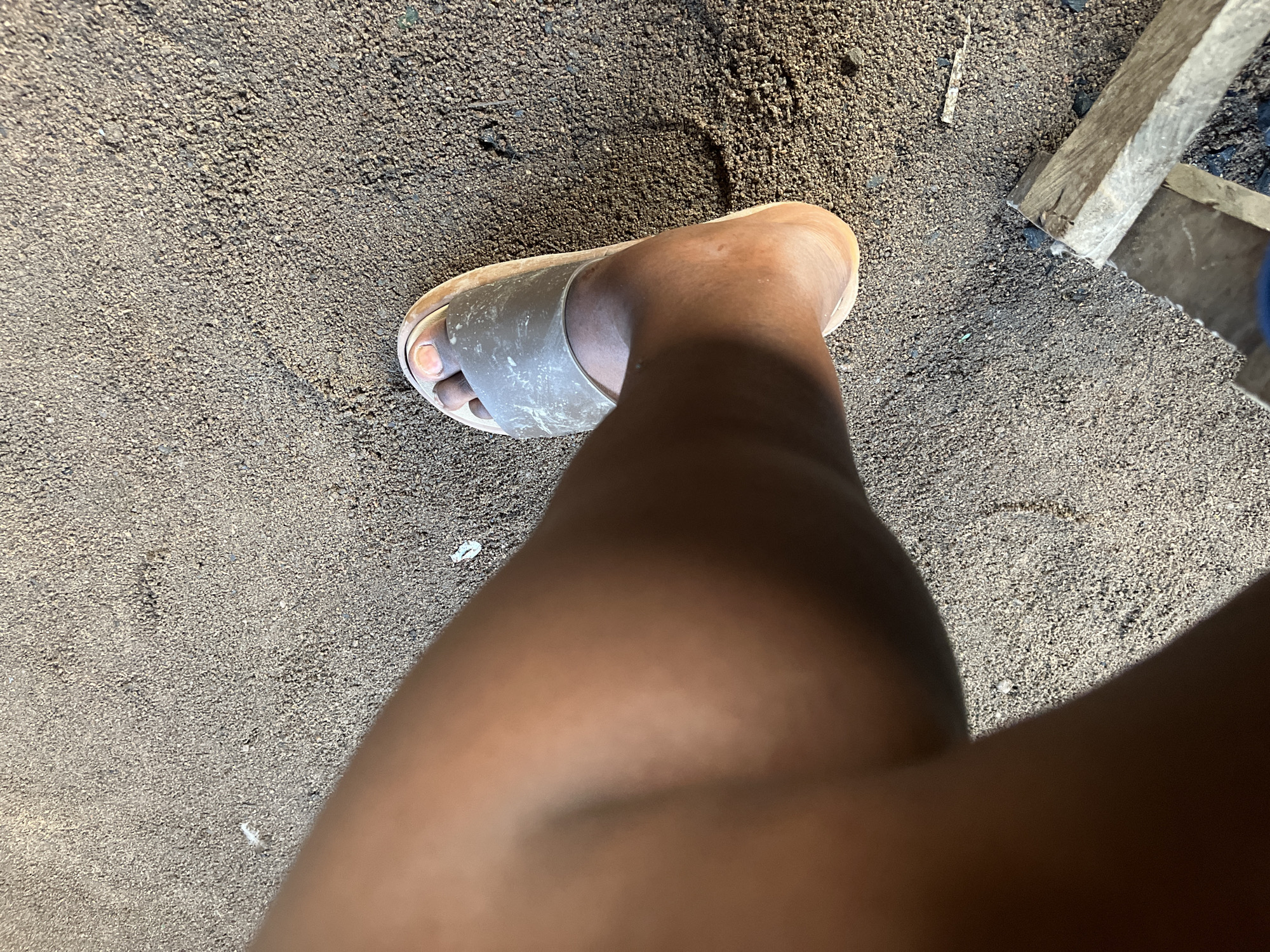 A person's left leg and foot are visible in the image, wearing a light-colored slide sandal. The leg is positioned at an angle, with the foot pointing towards the right side of the frame. The sandal is partially covering the foot, and the toes are visible with light-colored nail polish. The ground is covered in dark sand or dirt, with some small white specks that could be debris. In the lower right corner, two pieces of light-colored wood are visible, stacked on top of each other. The scene appears to be outdoors, possibly in a residential area or a dusty, unpaved space in Jalingo, Nigeria.