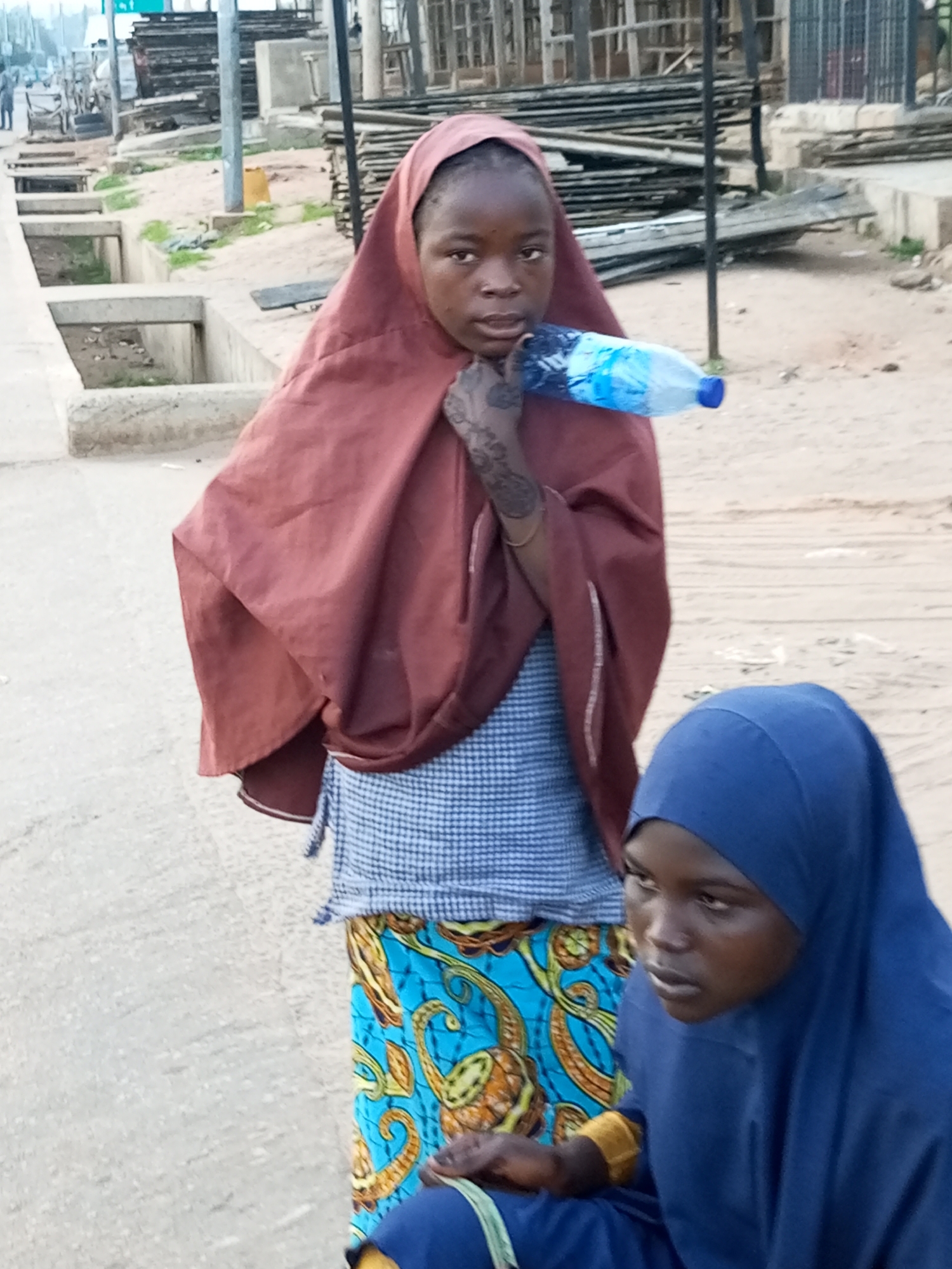 Two young girls are standing on a dirt and paved road in Gombe, Nigeria. The girl on the left is wearing a dark red headscarf, a blue and white checkered top, and a brightly patterned blue and orange skirt. She is holding a partially full plastic water bottle to her mouth. Her left hand, visible to the left of the bottle, has intricate henna or temporary tattoos. A gold bracelet is visible on her wrist. The girl on the right is wearing a royal blue headscarf and what appears to be a matching blue garment. She is looking down and to her right. Behind the girls, there are stacks of wood, some concrete drainage structures, and what appears to be a partially constructed building or scaffolding. The ground is a mix of dirt and paved areas. The sky is overcast.