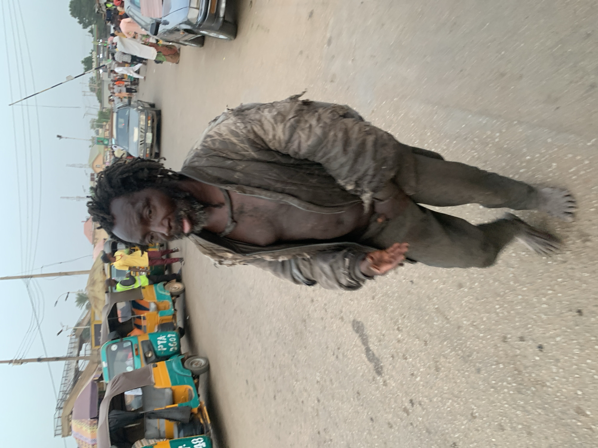 A side-angled shot depicts a dark-skinned man with dreadlocks and a beard standing on a paved street in what appears to be an urban area of Jos, Nigeria. He is the central and dominant figure in the foreground, facing slightly towards the left. His expression is dazed or distressed, with his eyes open but unfocused, and his tongue slightly extended from his mouth.
The man is clad in a torn, dirty, dark-colored jacket, worn open to reveal his bare chest, and matching dirty trousers. He is barefoot, and his feet appear grey with dust or dirt. His posture is hunched and leaning forward, with his hands raised, palms open, as if balancing or gesturing.
The background reveals a bustling street scene. To the man's left, a row of colorful auto-rickshaws, commonly known as keke-napep in Nigeria, are parked or moving slowly. Some passengers or drivers are visible near these vehicles. On one auto-rickshaw, the text "PTA 3602" is clearly visible, with partial text like "PTA" and "35." seen on others. To the man's right, older model cars are parked along the roadside, and more people can be seen in the distance, alongside buildings or market stalls. Overhead, power lines and a tall, slender pole extend into the hazy, overcast sky, suggesting daytime with muted light. The overall environment suggests a lively, perhaps crowded, street setting.