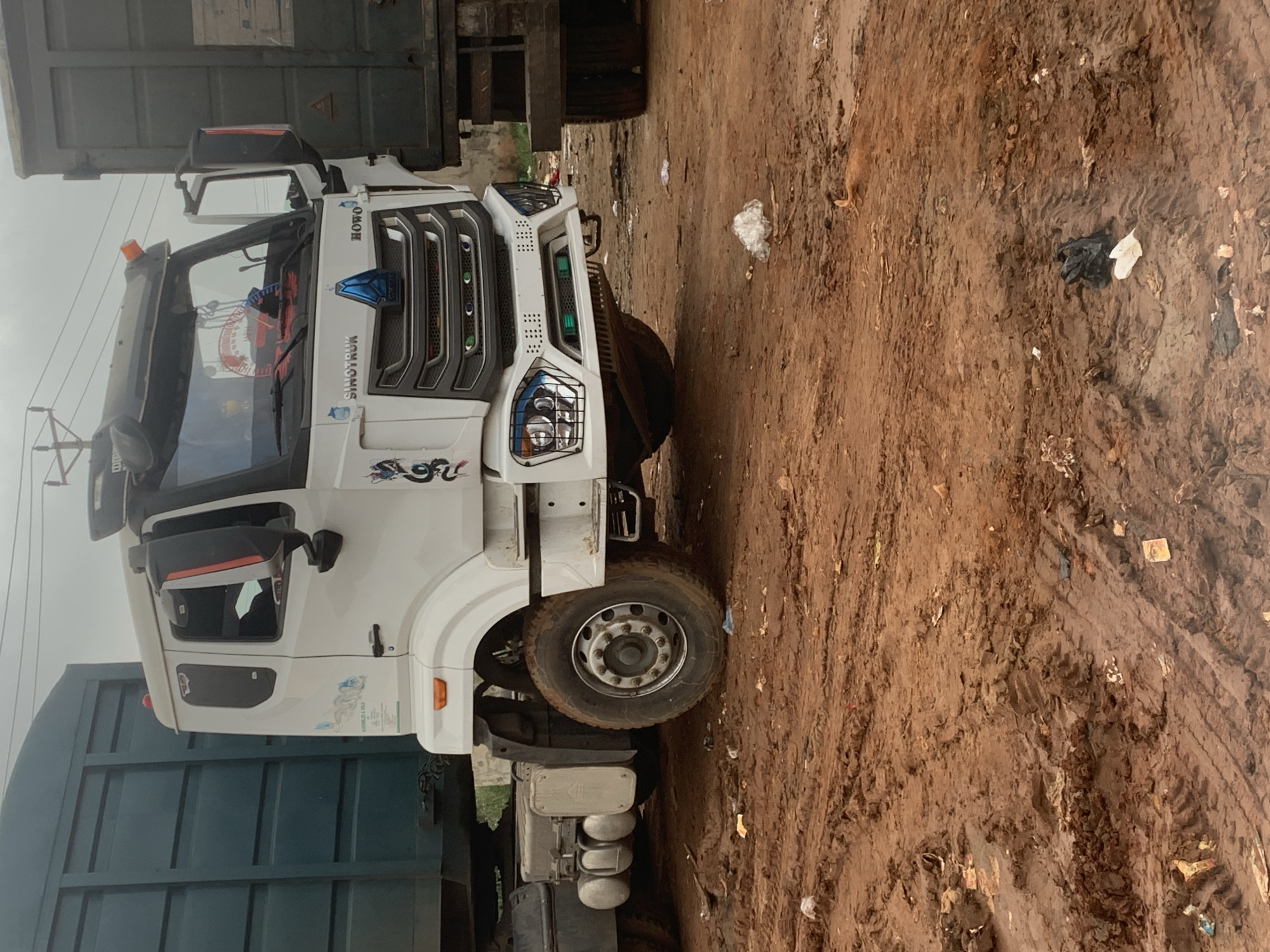 A white Sinotruk HOWO semi-truck is parked on a muddy, dirt lot in Shagamu, Nigeria. The truck's headlights are on and it is facing forward. There are other trucks parked around it, one to the left is dark teal and another to the right is a dark grey. The ground is covered in tire tracks and debris. The sky is overcast.