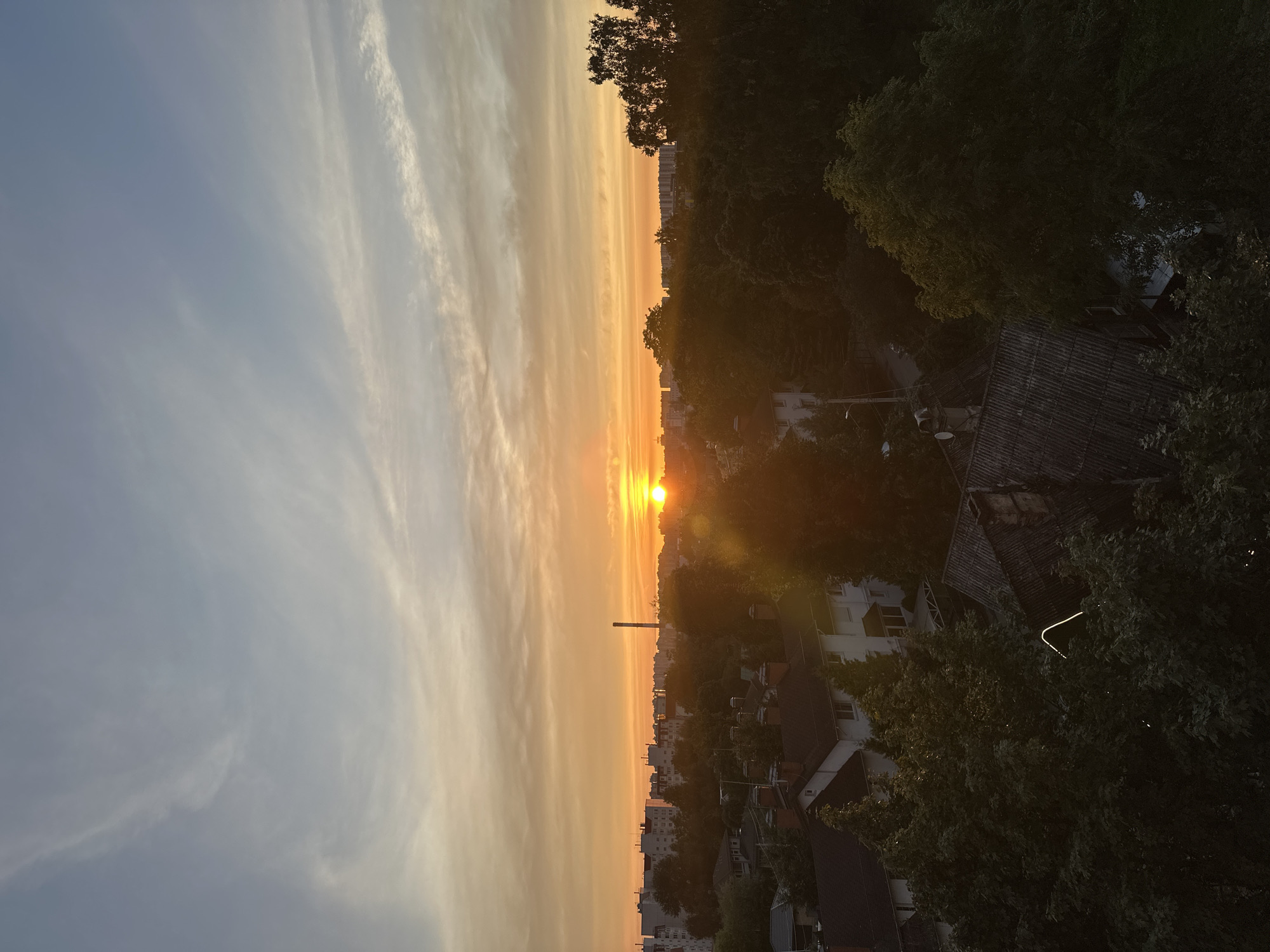 An aerial view captures a sunset over a residential area in Saint Petersburg, Russia. The sky is a gradient of soft blues and oranges, with wispy clouds streaking across the horizon. The sun, a bright orb, is setting just above the tree line, casting a warm glow over the scene. In the foreground, the dark silhouettes of trees obscure some of the buildings. These buildings appear to be a mix of houses with pitched roofs and taller apartment blocks in the distance. A tall, slender smokestack is visible in the middle distance. The overall impression is one of urban development intertwined with natural elements, illuminated by the soft, fading light of day.