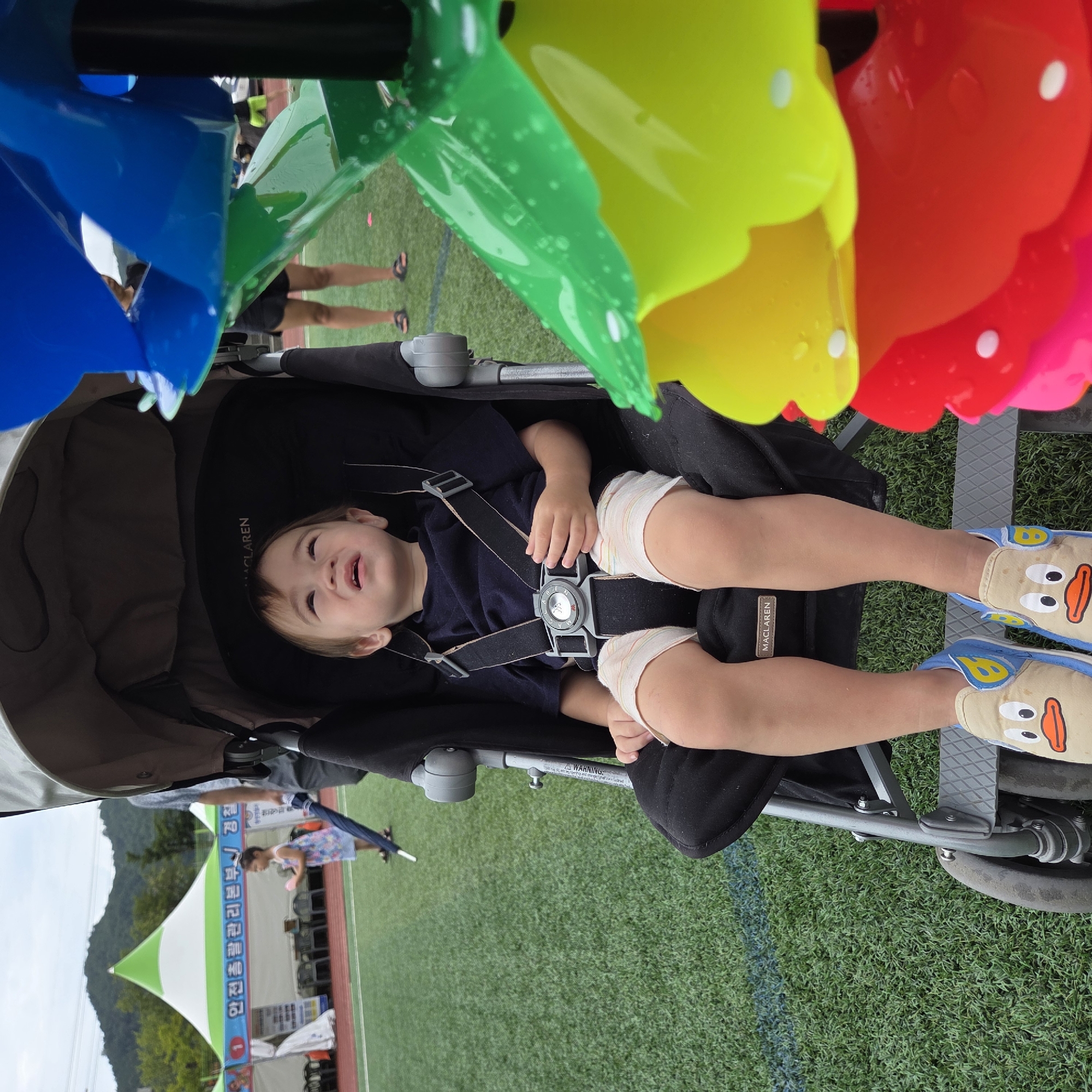 A young child sits in a black stroller on a green turf field. The child is wearing a dark t-shirt and light shorts with rainbow stripes. The child's feet are adorned with light brown slip-on shoes featuring cartoon duck faces. A large, colorful pinwheel toy is positioned to the right of the stroller, partially obscuring the child. In the background, a white and green tent is visible, with people standing nearby. The scene appears to be outdoors, possibly at an event or park. The weather is overcast, with a mountainous landscape visible in the distance. This scene is located in Dunnae-myeon, South Korea.