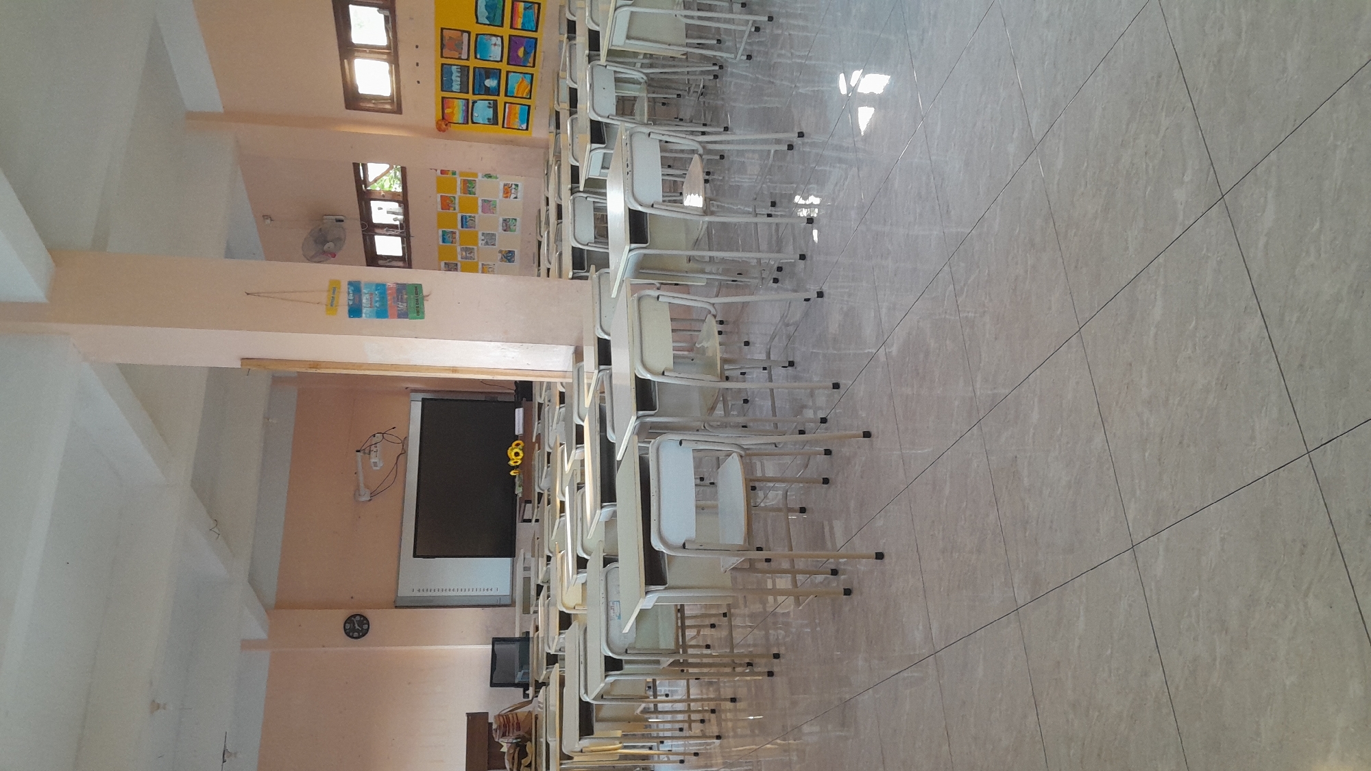 The image shows an empty classroom with rows of desks and chairs. The walls are painted a light peach color. A large interactive whiteboard is mounted on the front wall, with a projector above it. There are also some framed pictures and a fan on the wall. The floor is tiled with large square tiles. The room appears to be well-lit, with sunlight streaming in from windows. There are no people visible in the image. The scene is a typical classroom setup, likely for elementary or middle school students, in Tasikmalaya, Indonesia.