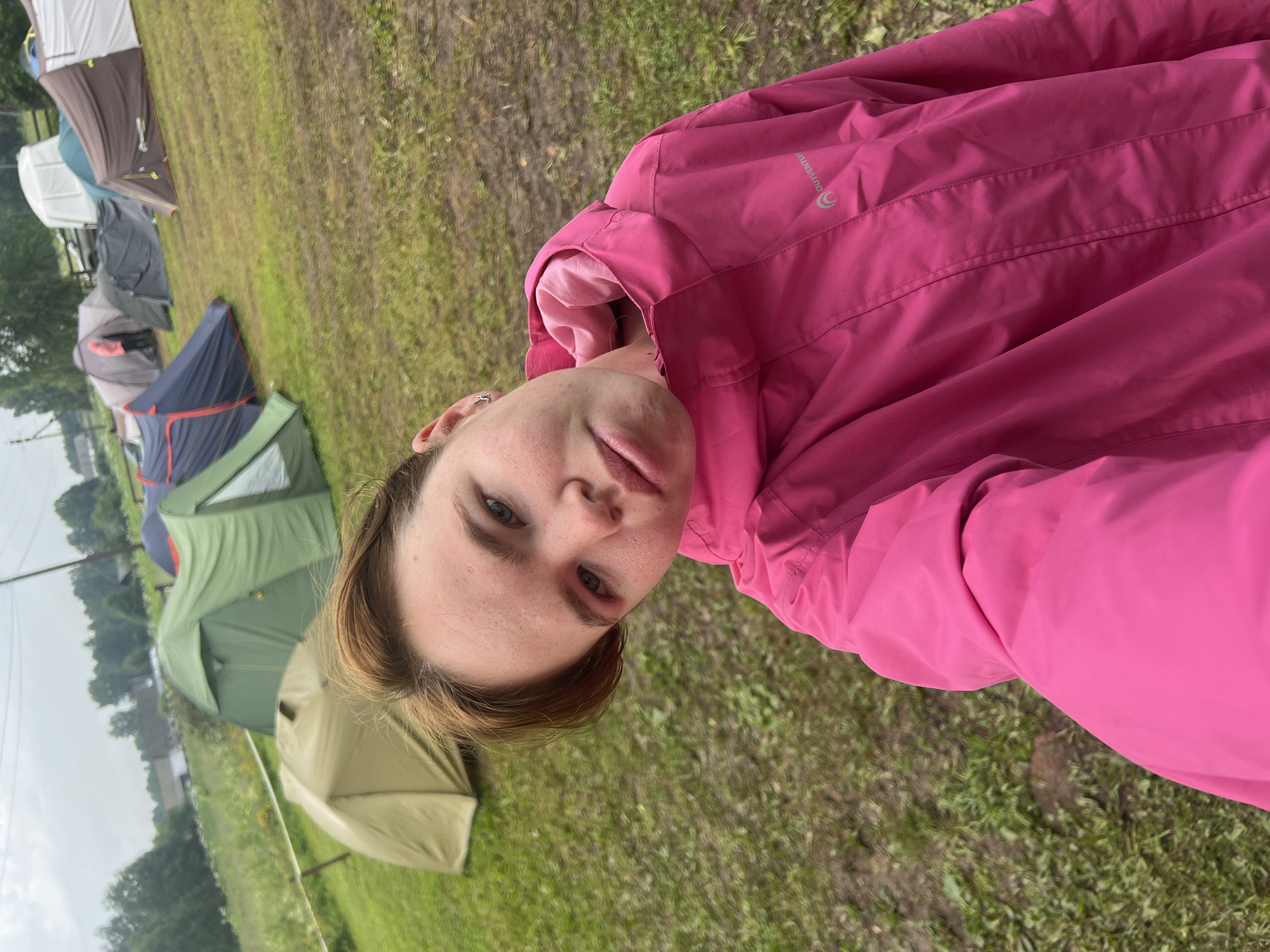 A medium shot, outdoor, slightly overcast day, selfie of a young Caucasian female wearing a bright pink hooded jacket. The woman has short, light brown hair and fair skin with some freckles. She is looking at the camera with a neutral expression, her head tilted slightly to the left. Her right eye appears to have pink eyeshadow. In the background, a grassy field is dotted with numerous tents of various colors including green, blue, and brown. Some trees and a cloudy sky are visible in the distance, suggesting a rural or camping environment. The overall impression is a candid moment captured during an outdoor activity, possibly a festival or camping trip.