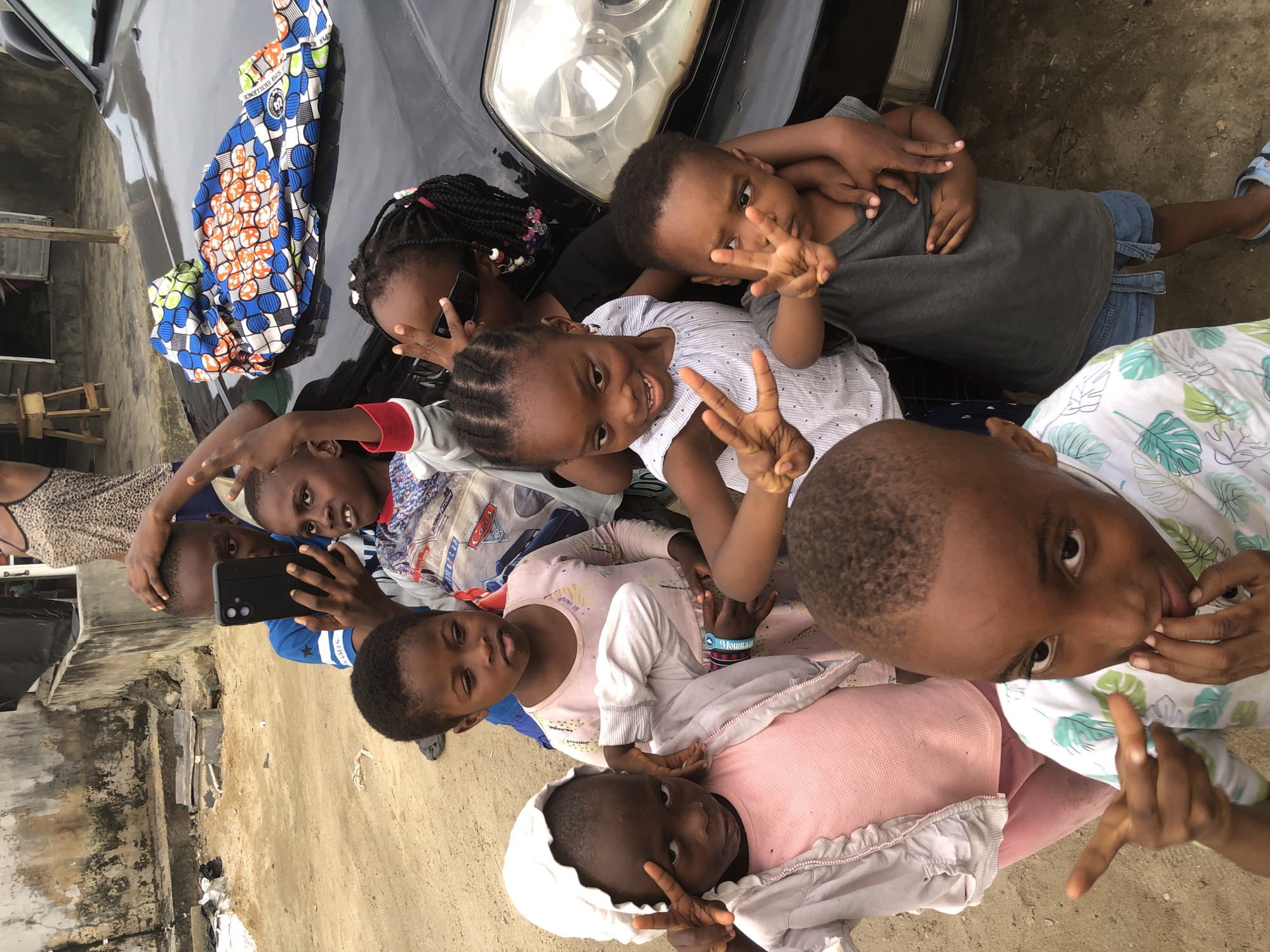 In the city of Ado, Nigeria, a group of Black children are gathered outdoors, with a black car as the backdrop. One child in the front left, wearing a pink top and a light pink hooded jacket, faces the camera and makes a peace sign with their right hand. Behind them, another child in a blue and white striped shirt, also makes a peace sign. A third child, in a dark gray t-shirt, stands beside them, also making a peace sign. In the background, an older child is holding a smartphone, seemingly taking a picture, and another child wearing a patterned blue, orange, and white fabric is draped over the car. The environment appears to be a sandy area with a textured, weathered building in the background, suggesting a residential or informal settlement.